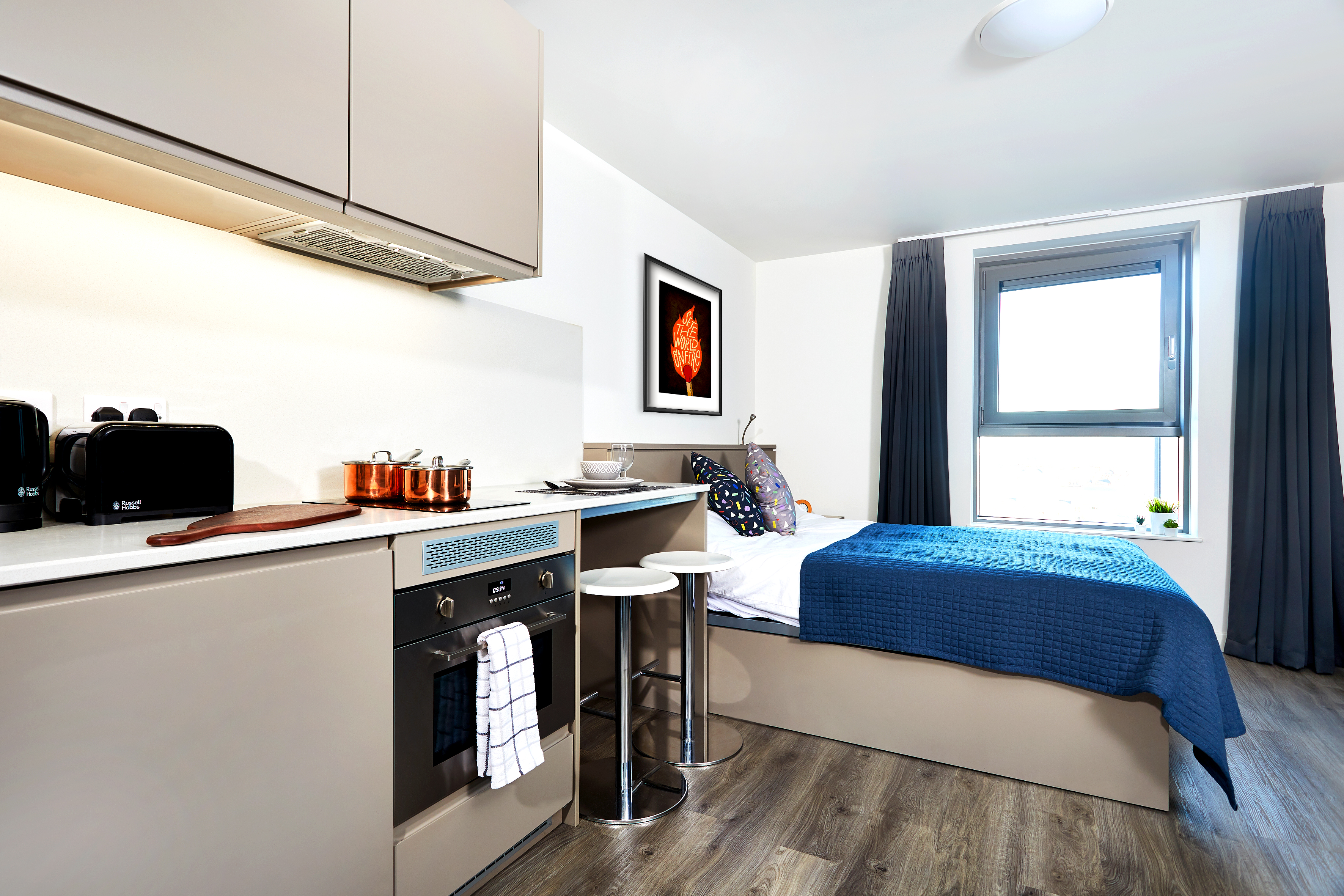 Studio apartment with a compact kitchen, featuring beige cabinets, an oven, and a toaster. A round table with two stools is nearby. The bed has a blue quilt. A window with curtains and a framed artwork on the wall complete the cozy setting.
