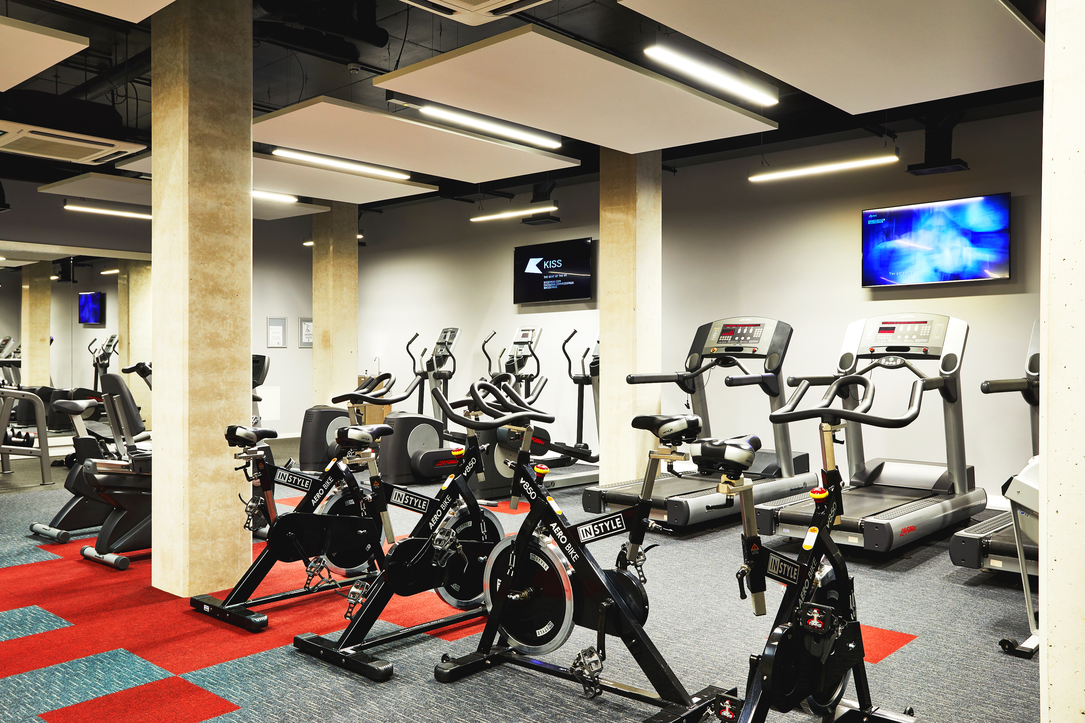 A modern gym with exercise bikes, treadmills, and a few flat-screen TVs mounted on the walls. The room has a clean design with gray walls, light wood pillars, and red and gray flooring.