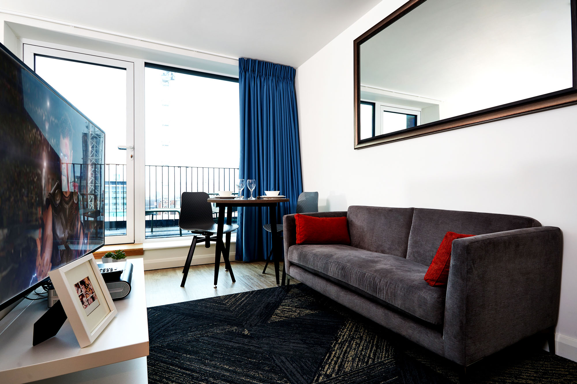 Modern living room featuring a gray sofa with red cushions and a large square wall mirror, complemented by a flat-screen TV on a stand. A small dining table with chairs sits by the window with blue curtains, leading to a balcony that frames the city like an enchanting urban circle.