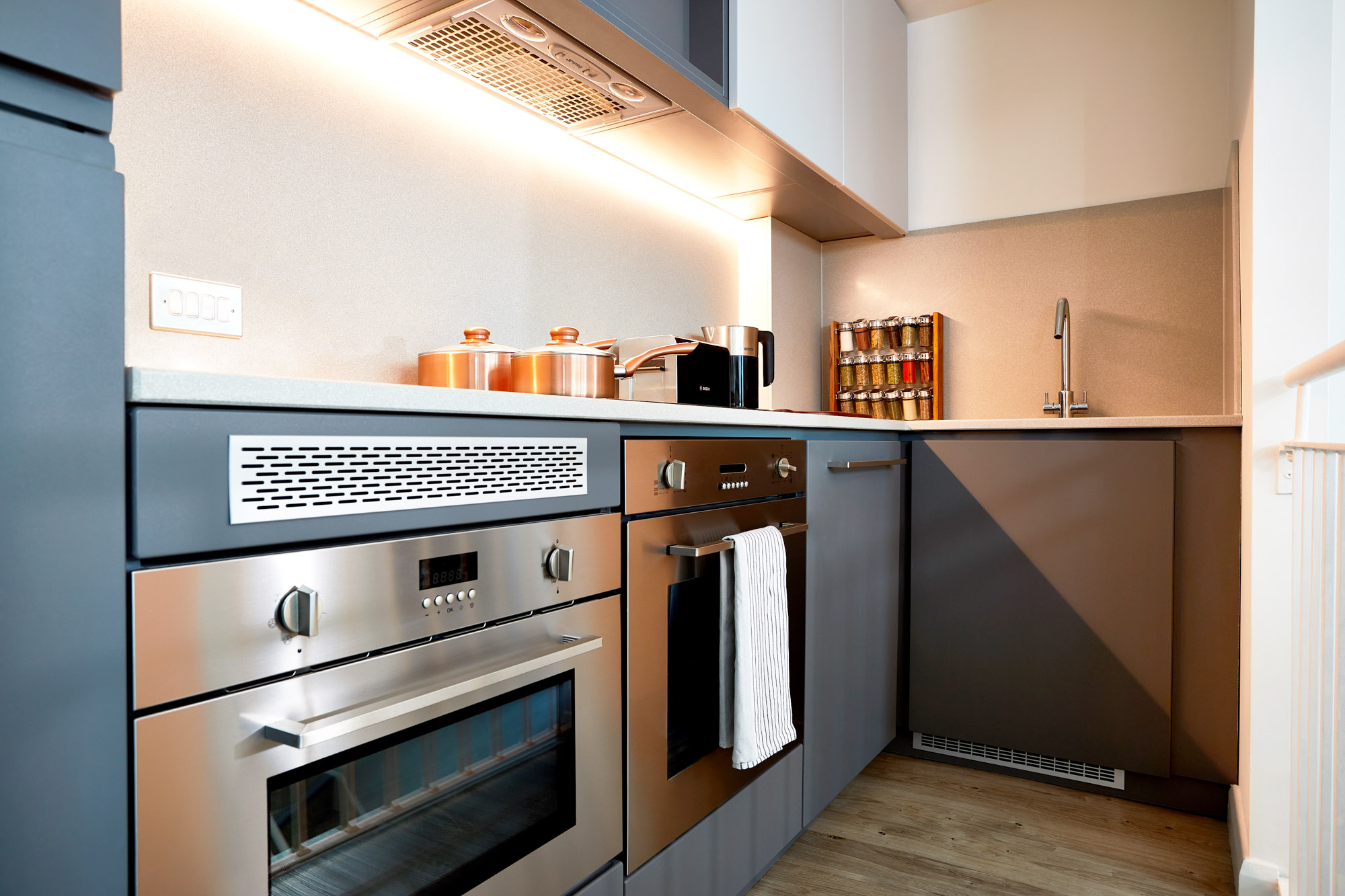Modern kitchen with stainless steel appliances, including an oven and range hood. Grey cabinets and a light countertop hold copper pots, a knife rack, and spice jars. Warm lighting under cabinets and a wooden floor complete the space.