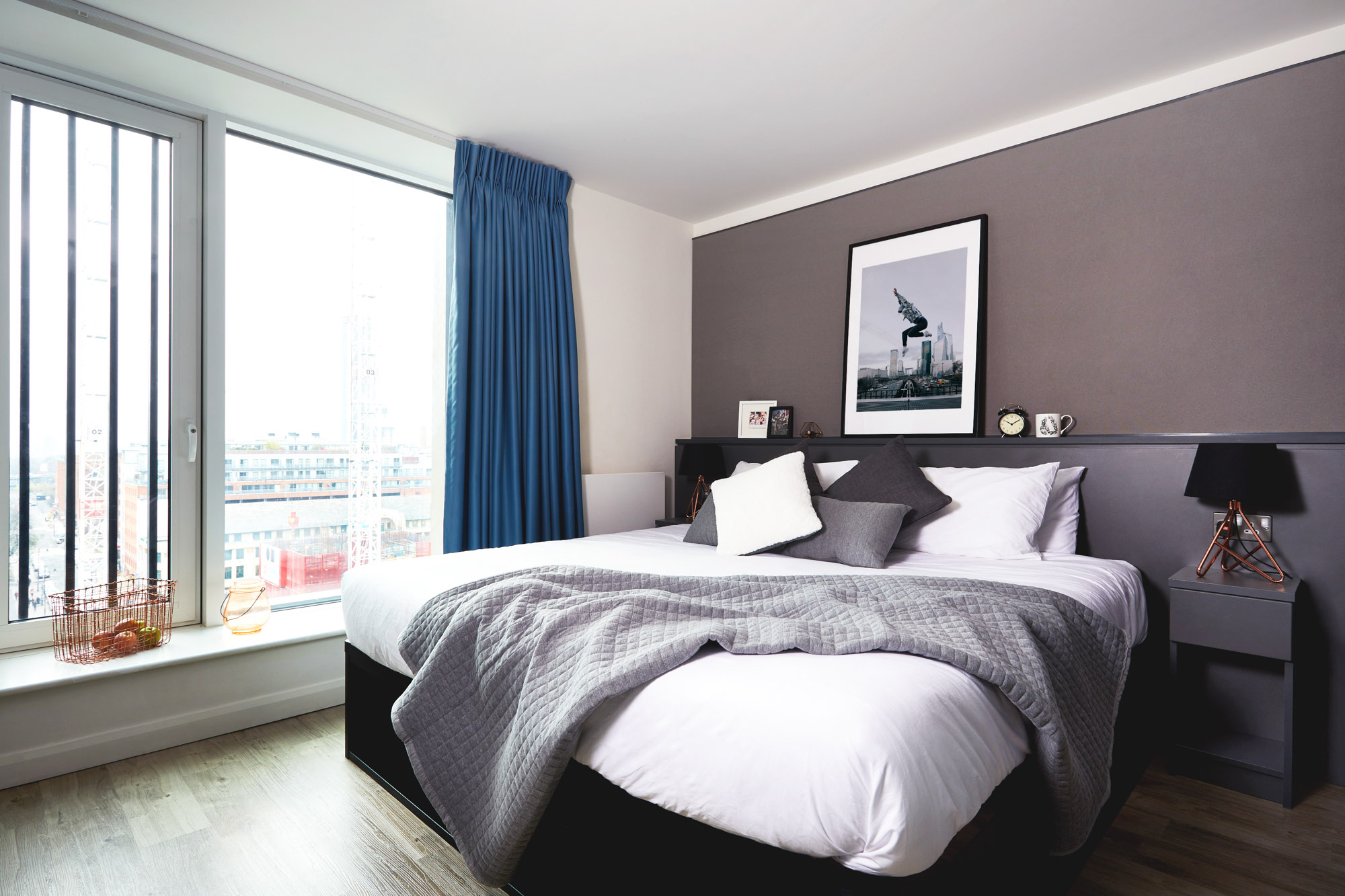 Modern bedroom with a large bed covered in gray and white bedding, a framed square artwork on the gray wall, and a side table with a circular black lamp. A floor-to-ceiling window with blue curtains offers city views. Neutral tones create a cozy ambiance.