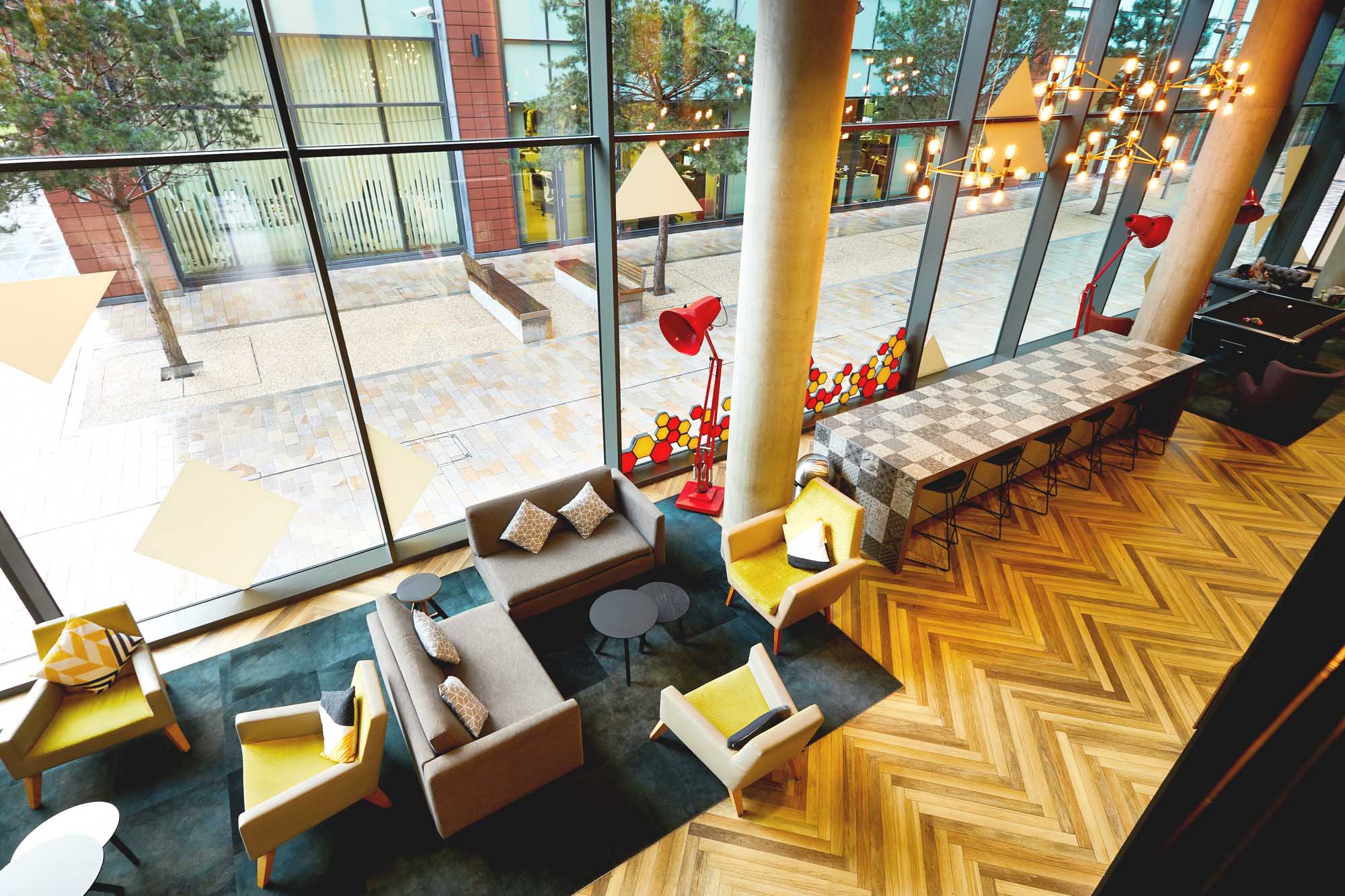 Modern lounge on First Street with large windows, wooden herringbone floor, and contemporary furniture. Features grey sofas, yellow chairs, small round tables, and red lamps. A tall counter with stools runs alongside the windows. Outside, trees and brick buildings are visible.