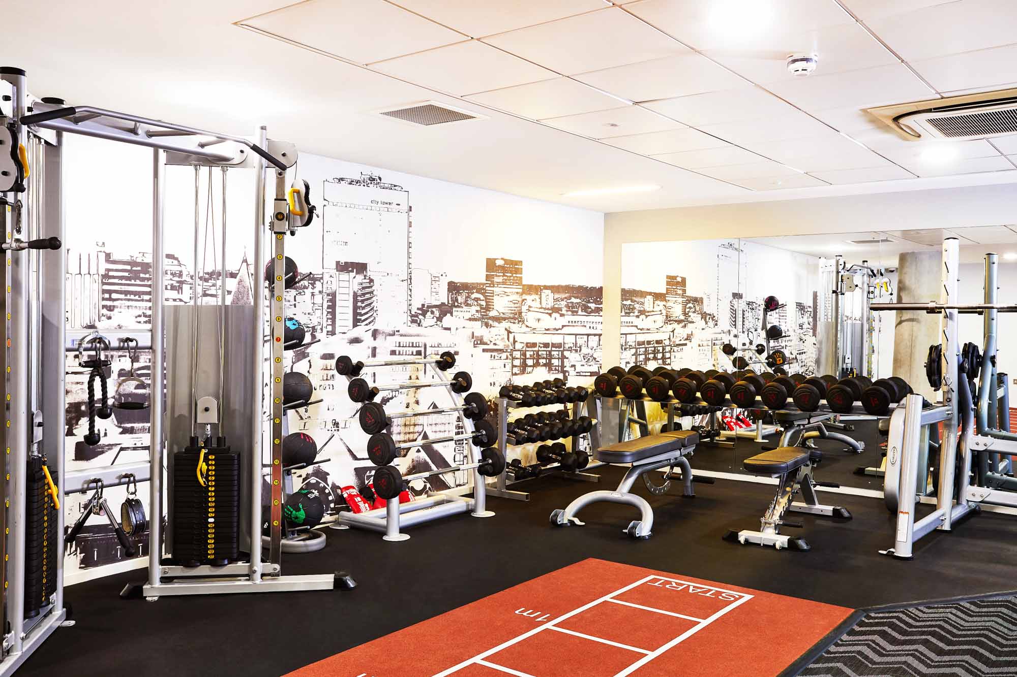 A modern gym with weightlifting equipment, including dumbbells, barbells, and benches. The room has wall art of a cityscape, mirrors, and an area with resistance machines. The floor has an exercise grid pattern.