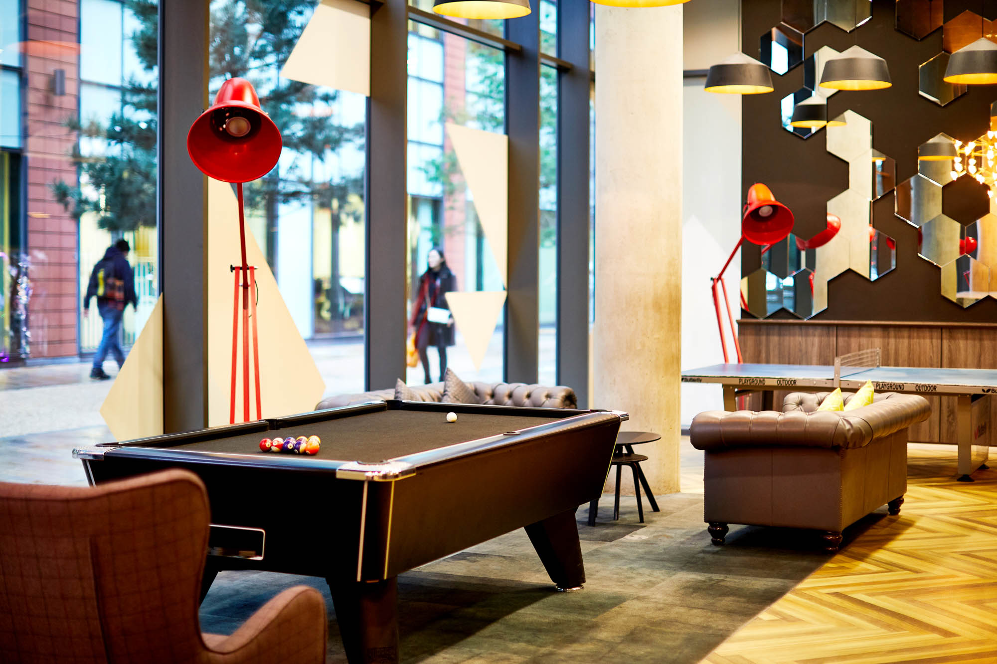 Located on First Street, a modern lounge boasts a black pool table, stylish brown sofas, and unique geometric wall mirrors. Large windows reveal people walking outside. Red floor lamps and wooden flooring add warmth to the contemporary space.