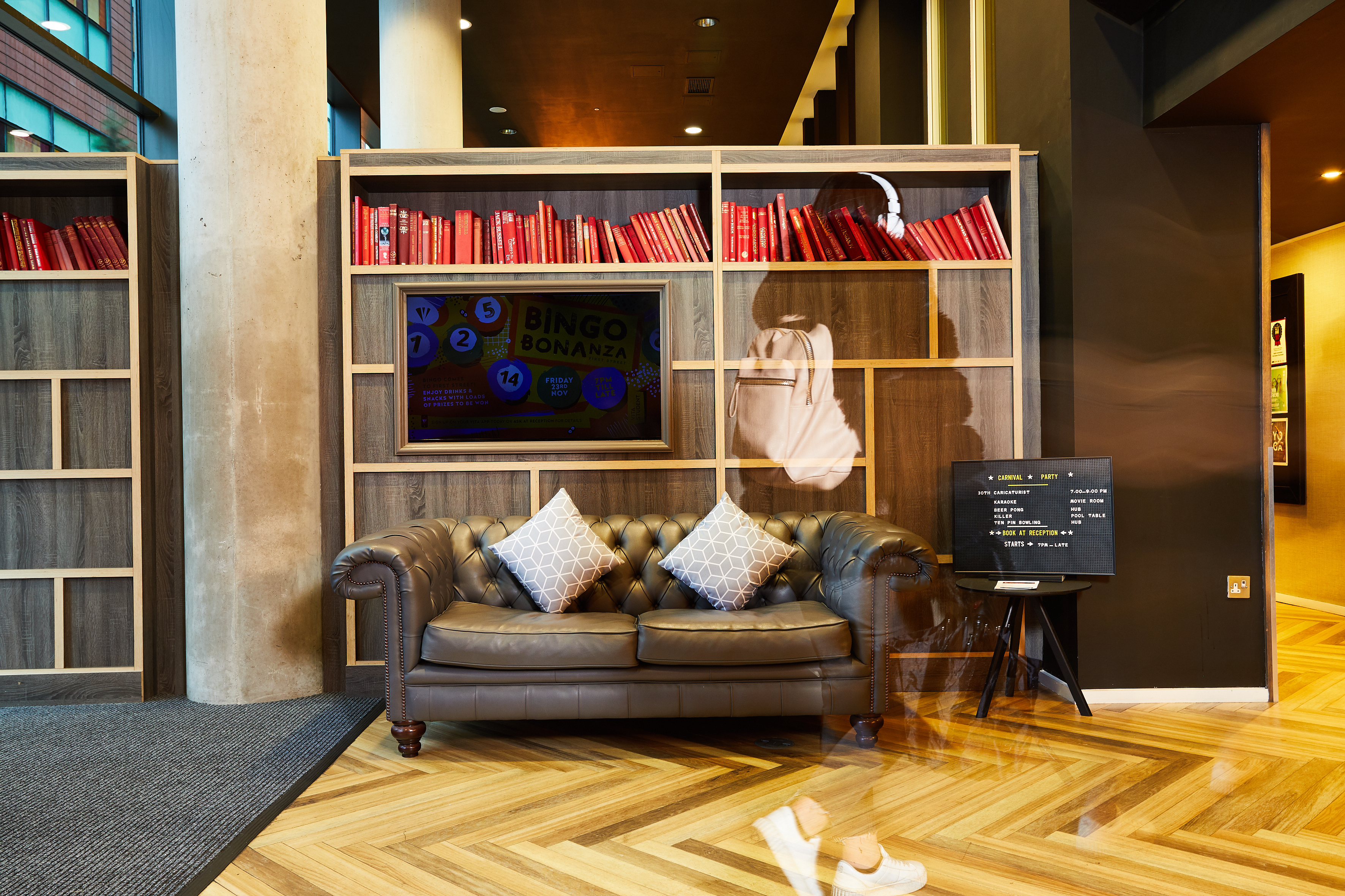 A modern lobby featuring a brown leather sofa with two patterned cushions, a bookshelf filled with red books, and a TV displaying "Bingo Bonanza." A blurred figure walks by, creating a motion effect on the wooden floor.