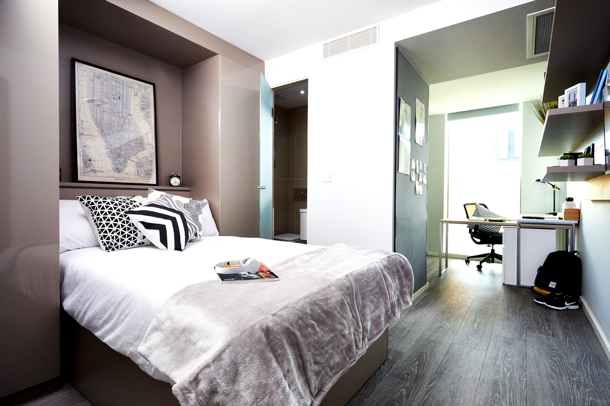 A modern bedroom on First Street features a neatly made bed, patterned pillows, and a soft throw blanket. A desk with a chair sits near the window, while shelves filled with books and decor line the wall. The space boasts a minimalist design with clean lines and neutral colors.