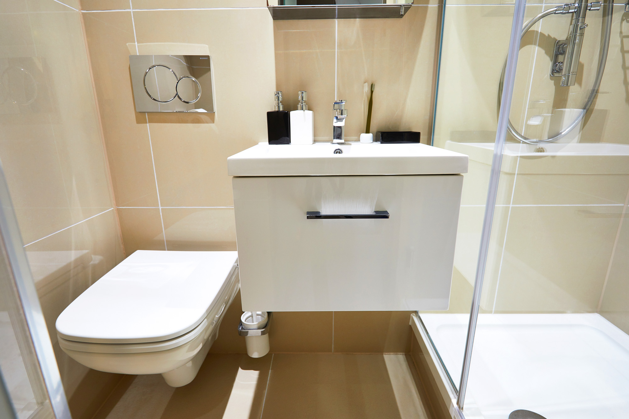 This modern bathroom on First Street features beige tiles, a floating toilet, a white wall-mounted sink with toiletries, and a glass-enclosed shower with chrome fixtures.