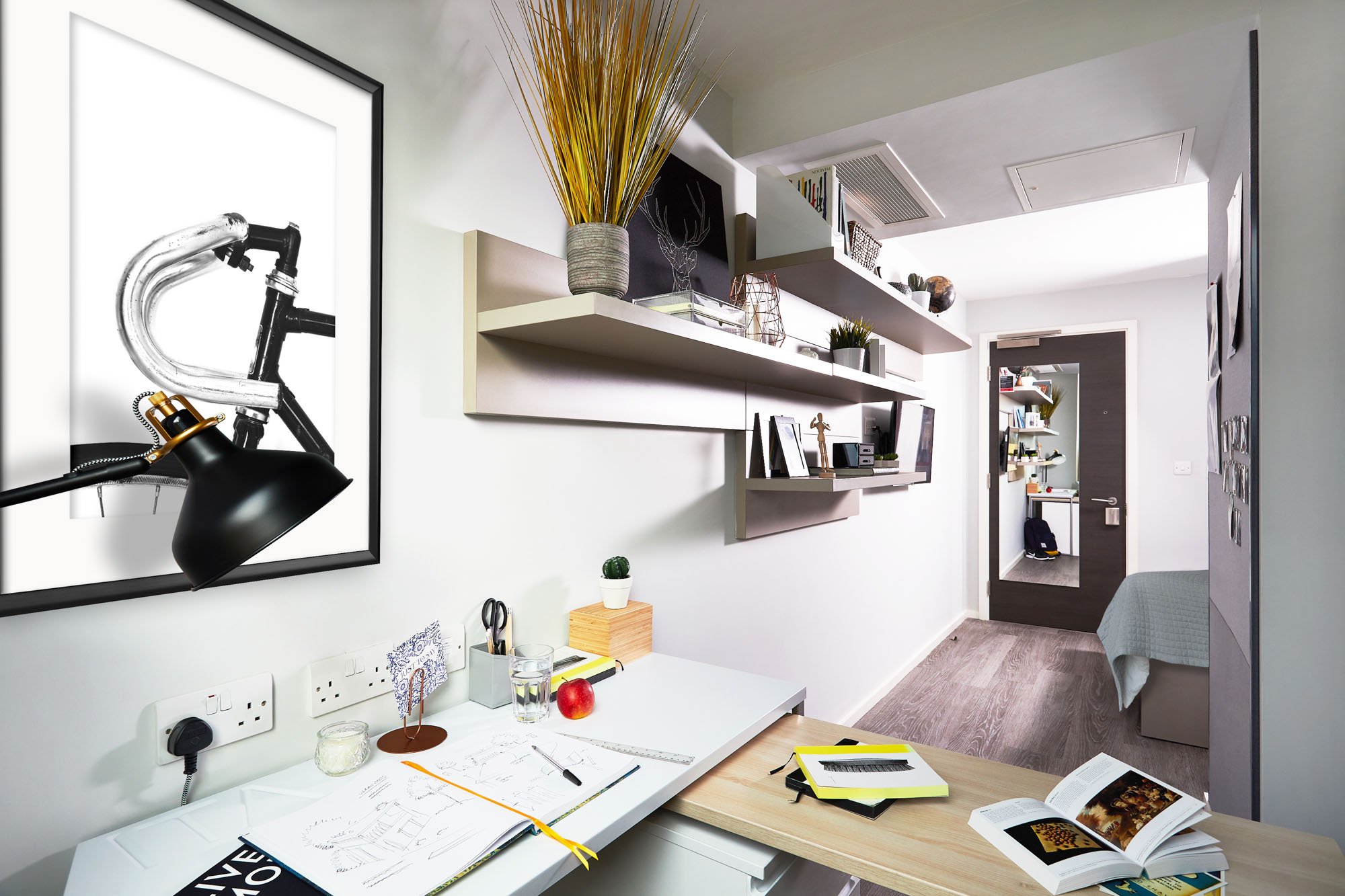 A modern, compact room on First Street boasts a desk adorned with books, a black lamp, an apple, and stationery. Shelves display decorative items. A bed is visible near a door against crisp white walls, beautifully offset by elegant wooden floors.