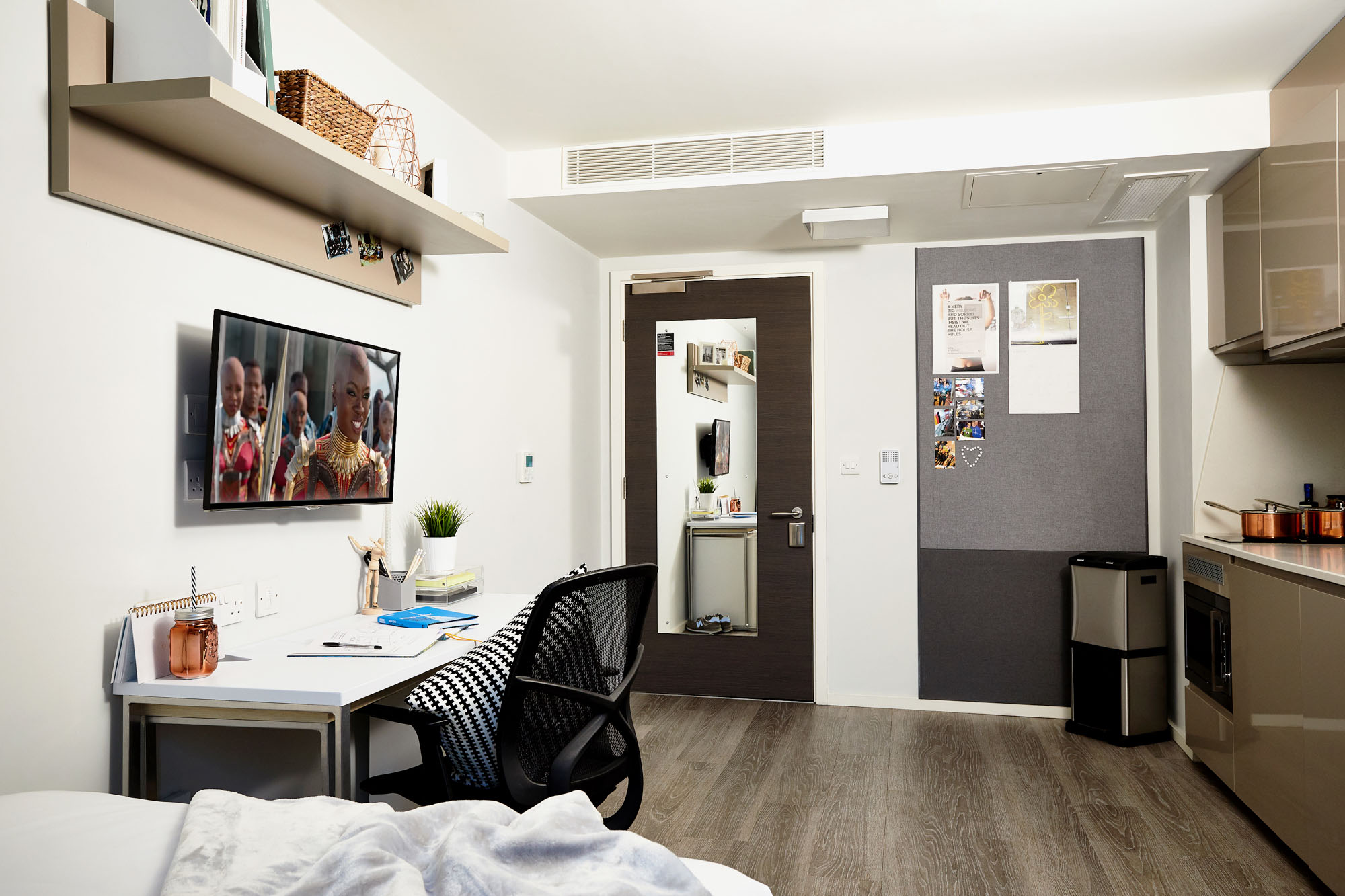 Located on First Street, this modern dorm room features a wall-mounted TV, a study desk with a chair, and stylish décor. It includes a cozy bed with a blanket, a kitchenette adorned with copper pots, and a mirrored door. Personal touches adorn the bulletin board.