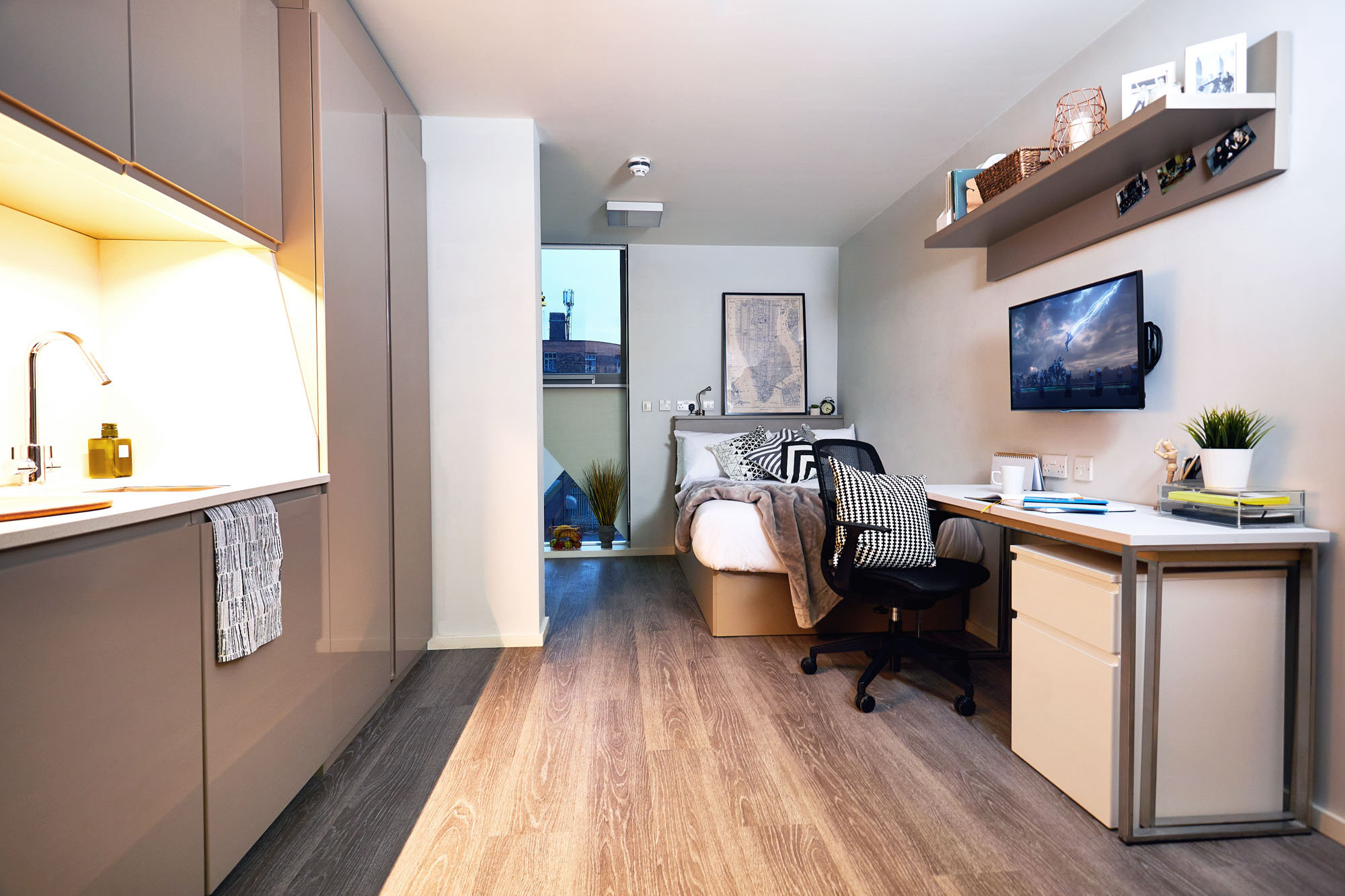 This modern student dorm room on First Street features a bed, desk, chair, and wall-mounted TV. The kitchenette with cabinets is on the left. Above the TV, shelving showcases plants and decor. A large window offers an urban view. Light-colored walls and wood flooring complete the inviting space.