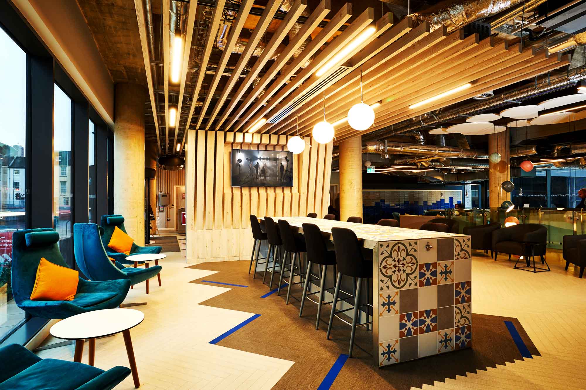 Modern office lounge with high ceiling, featuring a long table with patterned tiles and bar stools. Blue chairs with orange cushions line the window. Exposed ceiling, wooden slats, pendant lights, and a TV mounted on a partition wall enhance the decor.