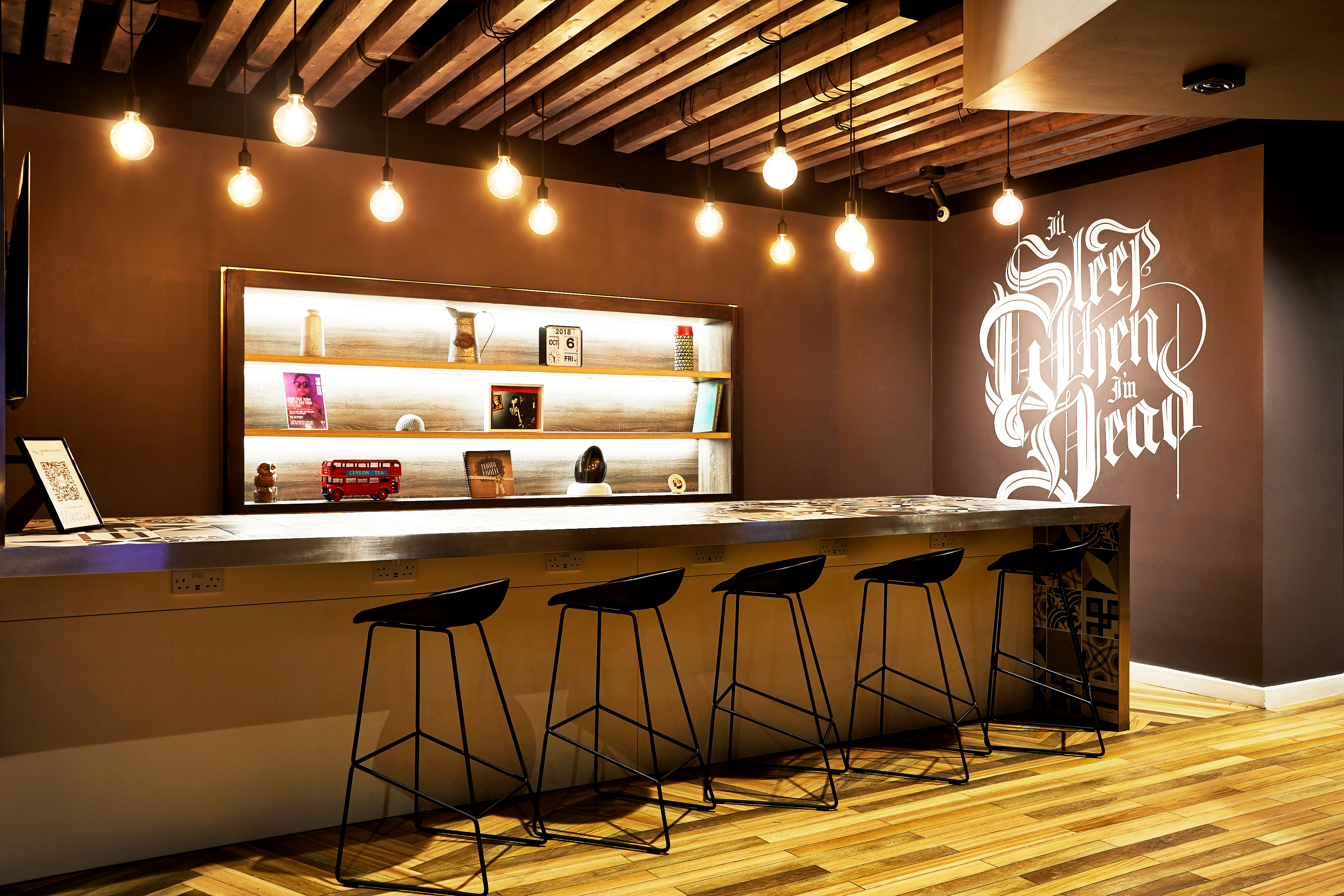 Modern cafe with wooden flooring and ceiling, dim overhead lights, and a long bar counter with black stools. Shelves display books and decor items. A wall features elegant white script text.
