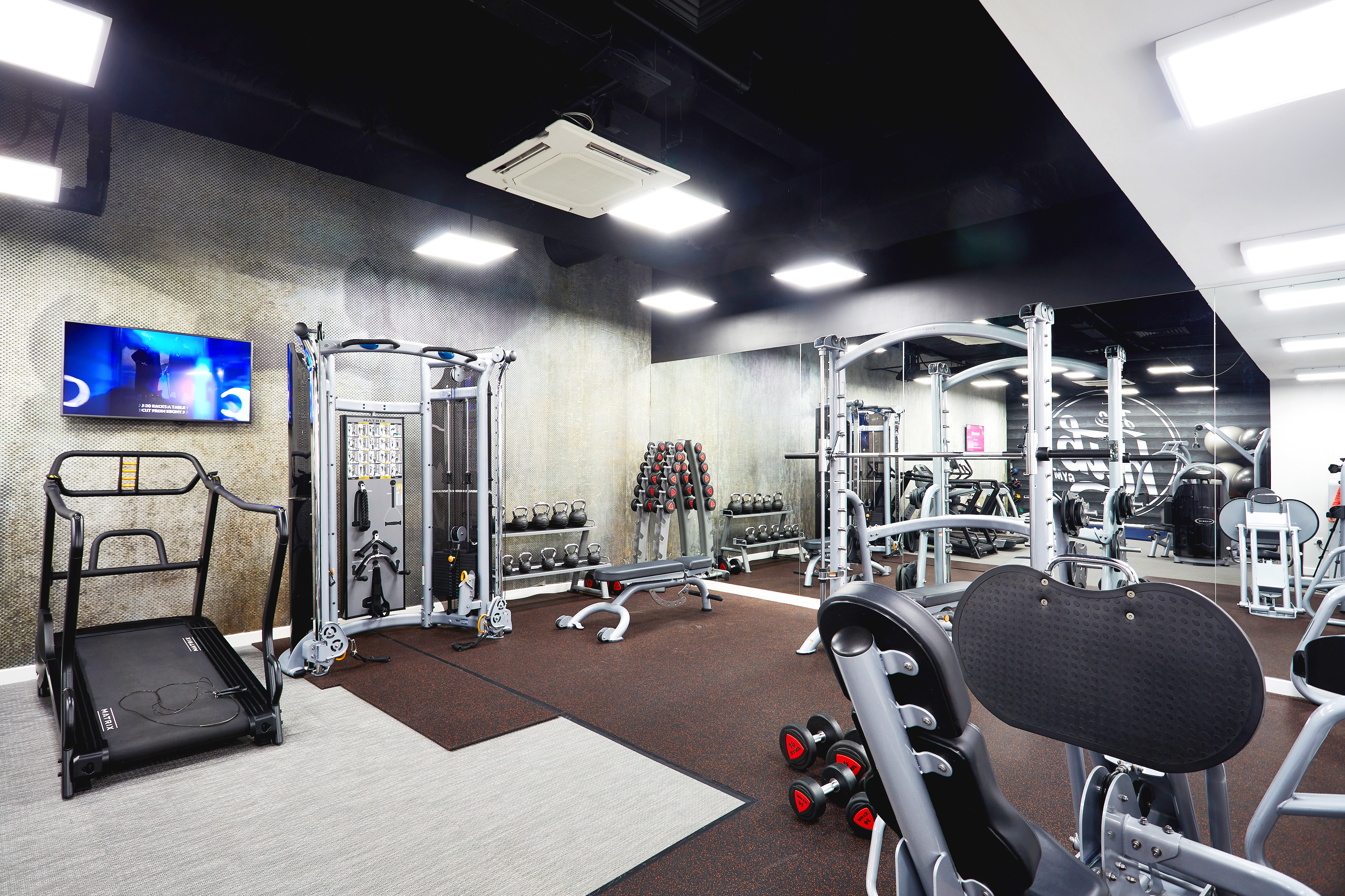 A modern gym with exercise equipment, including a treadmill, multi-use machine, free weights, and benches. The walls have large mirrors and there's a television screen on the left. Bright overhead lighting illuminates the space.