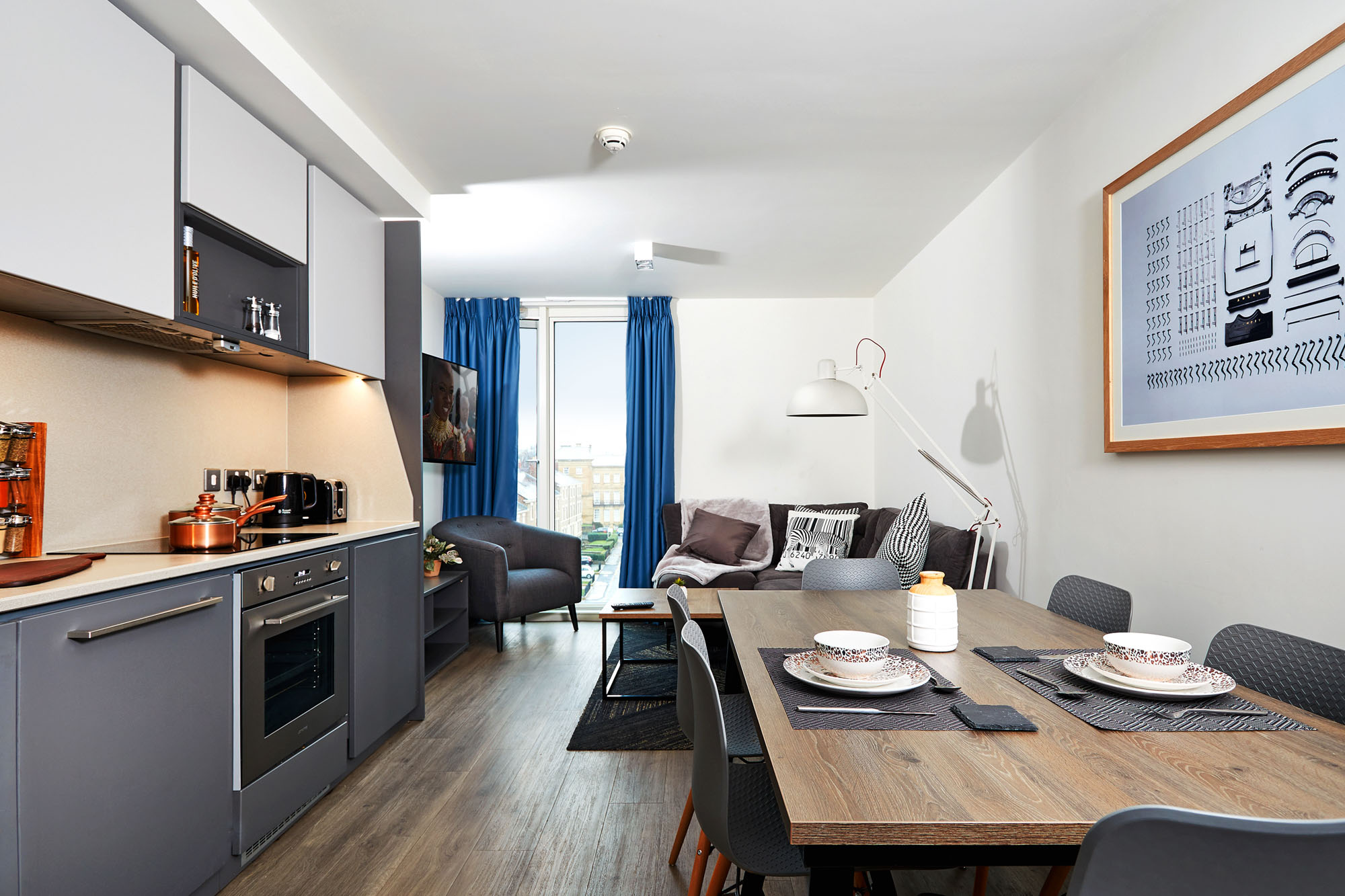 Modern apartment interior at Strawberry Place featuring a kitchen with gray cabinets, stove, and sink on the left. A dining table with place settings is in the foreground, while a cozy sitting area with chairs, a sofa, and TV is visible in the background. A large window allows light in.