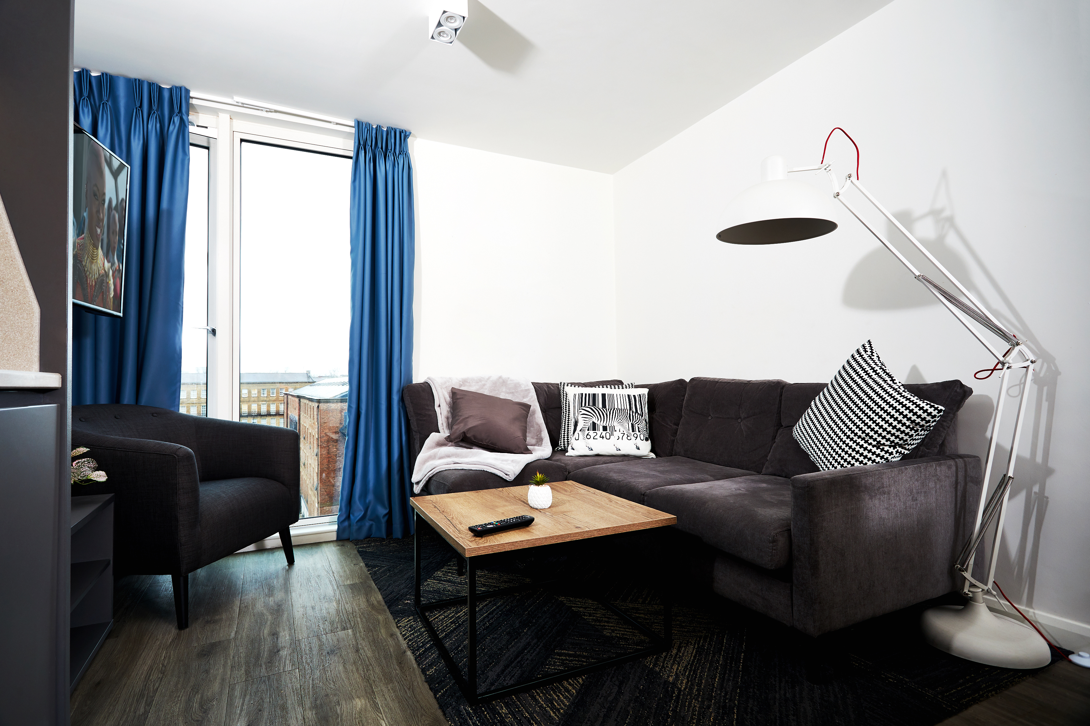 In the Strawberry Place living room, a dark gray sectional sofa with patterned pillows invites relaxation. A large white floor lamp gracefully arches over the couch, while a wooden coffee table holds a candle and remote. Blue curtains frame a tall window, accompanied by a dark armchair to the side.