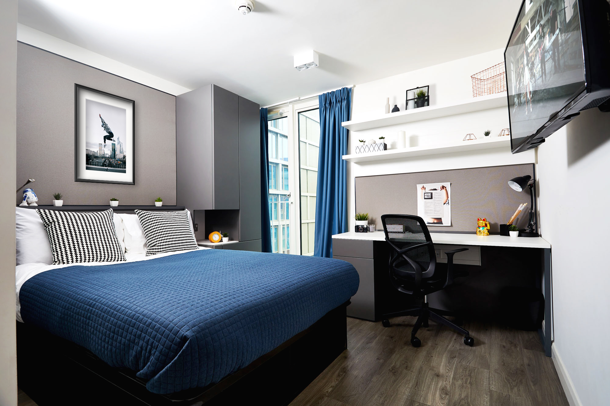 A modern bedroom with a grey and white color scheme features a bed adorned with a blue duvet and striped pillows, alongside shelves displaying decor and perhaps even a small strawberry plant. A desk with a chair faces large windows draped in blue curtains, allowing natural light to pour into the inviting place.