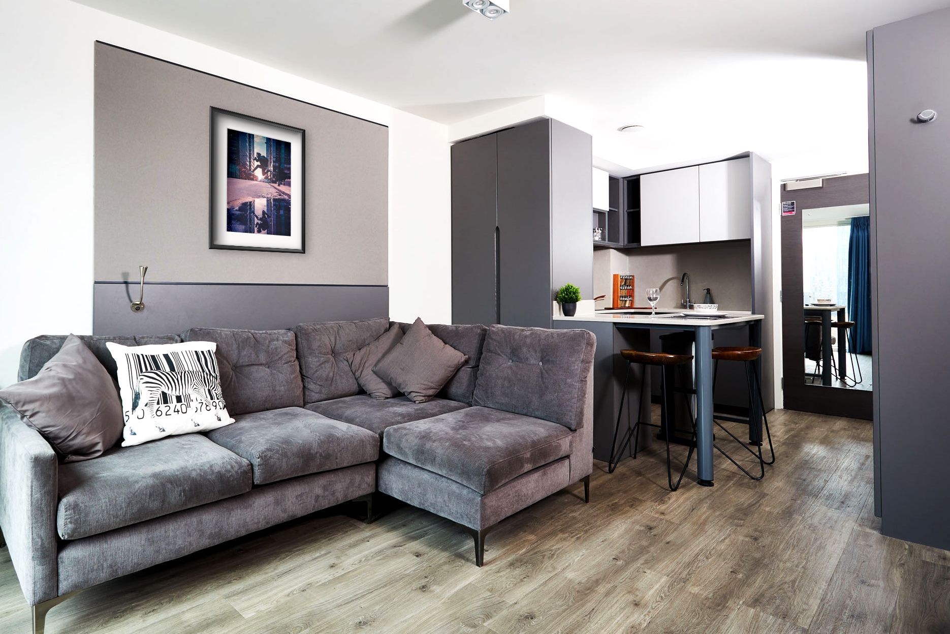 A modern living room at Strawberry Place features a gray L-shaped sofa with patterned cushions, facing a small kitchen. The kitchen includes a breakfast bar with stools. Artwork decorates the wall above the sofa, and the space boasts wooden flooring and gray cabinets.