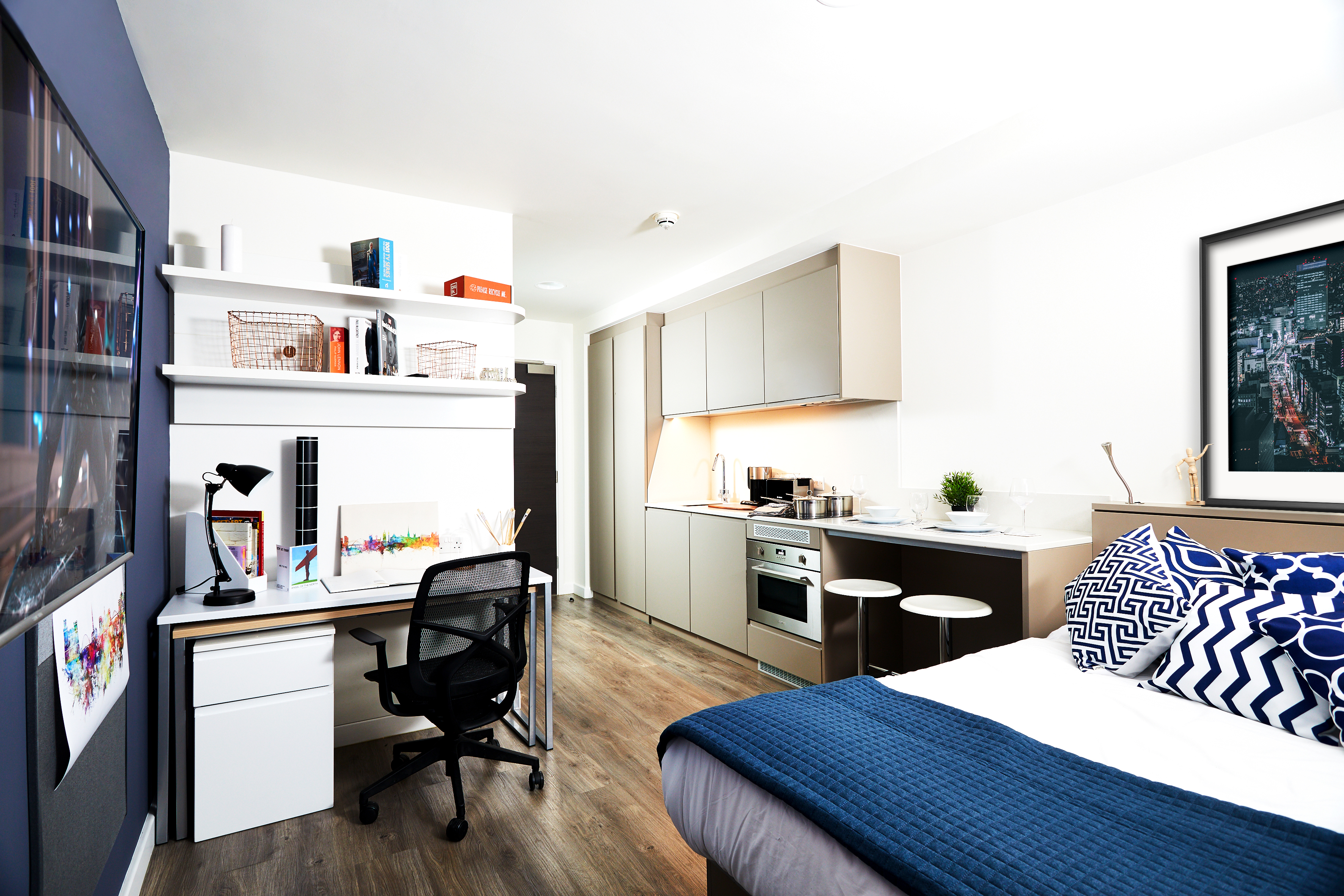 Modern studio apartment with a bed, desk, and kitchen. The bed has blue and white pillows and a navy throw. The desk includes a lamp and chair. Kitchen area has stools, shelves, and appliances. Walls feature artwork, and flooring is wood.