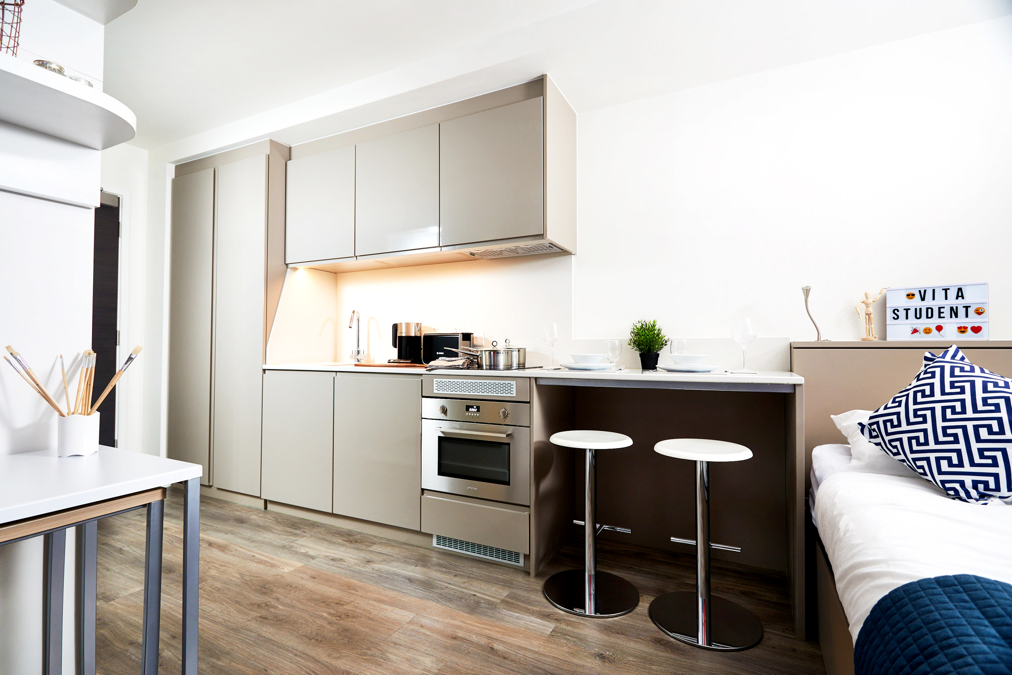 Modern kitchen with light brown cabinets and stainless steel appliances. A small bar with two stools is adjacent to a bed with white and blue bedding. A "Vita Student" sign and potted plant add decor. The floor is wood-style laminate.