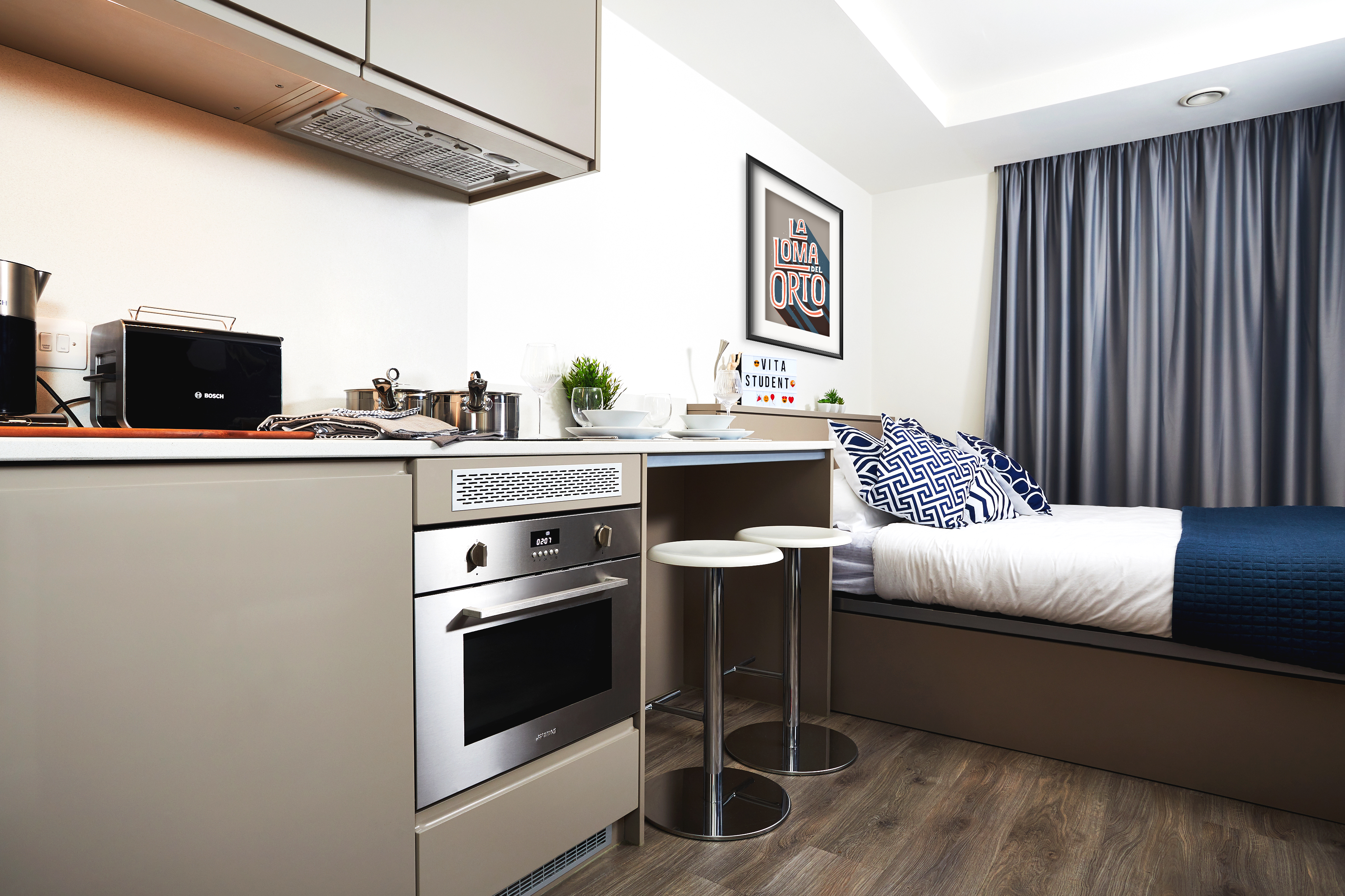 Compact modern studio apartment with a small kitchen featuring an oven, coffee maker, and toaster. A bed with geometric pillows is adjacent, and a counter with two stools sits nearby. A decorative print hangs on the wall above the bed.