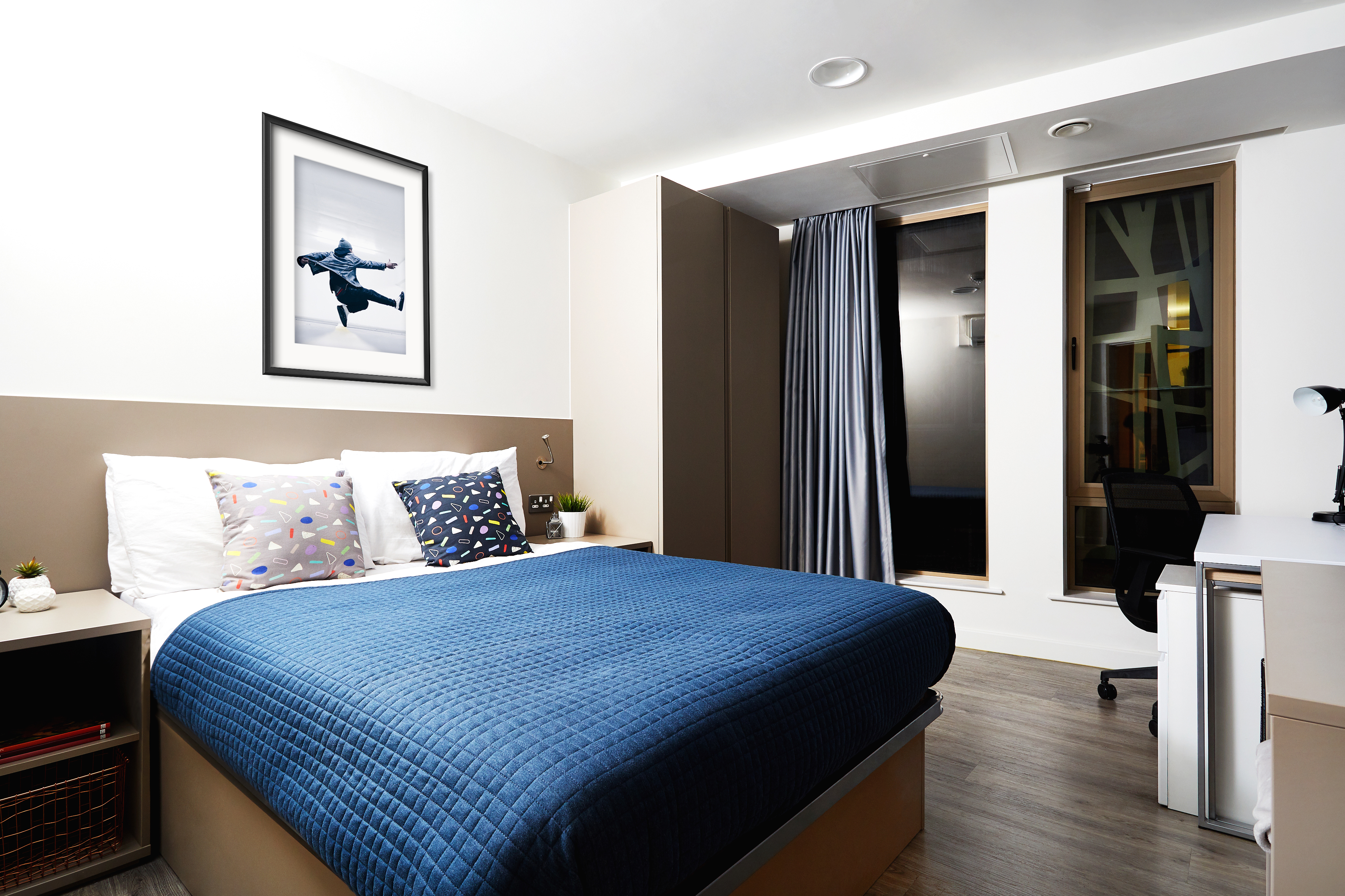 Modern bedroom with a blue quilted bed, colorful cushions, and a wall art piece featuring a plane. There's a desk with a chair and a lamp to the right, large windows with curtains, and light wood flooring. Neutral-toned walls complement the decor.