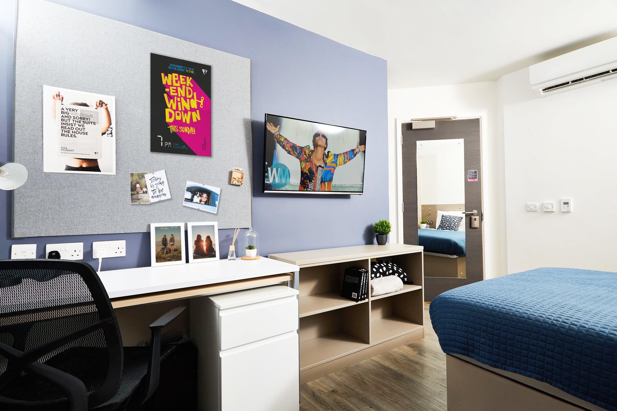 A modern dorm room with a desk, chair, and wall-mounted TV. Posters and photos decorate the walls. A bed with blue bedding is visible. The room is brightly lit, featuring a mirror on the door and sleek storage shelves.