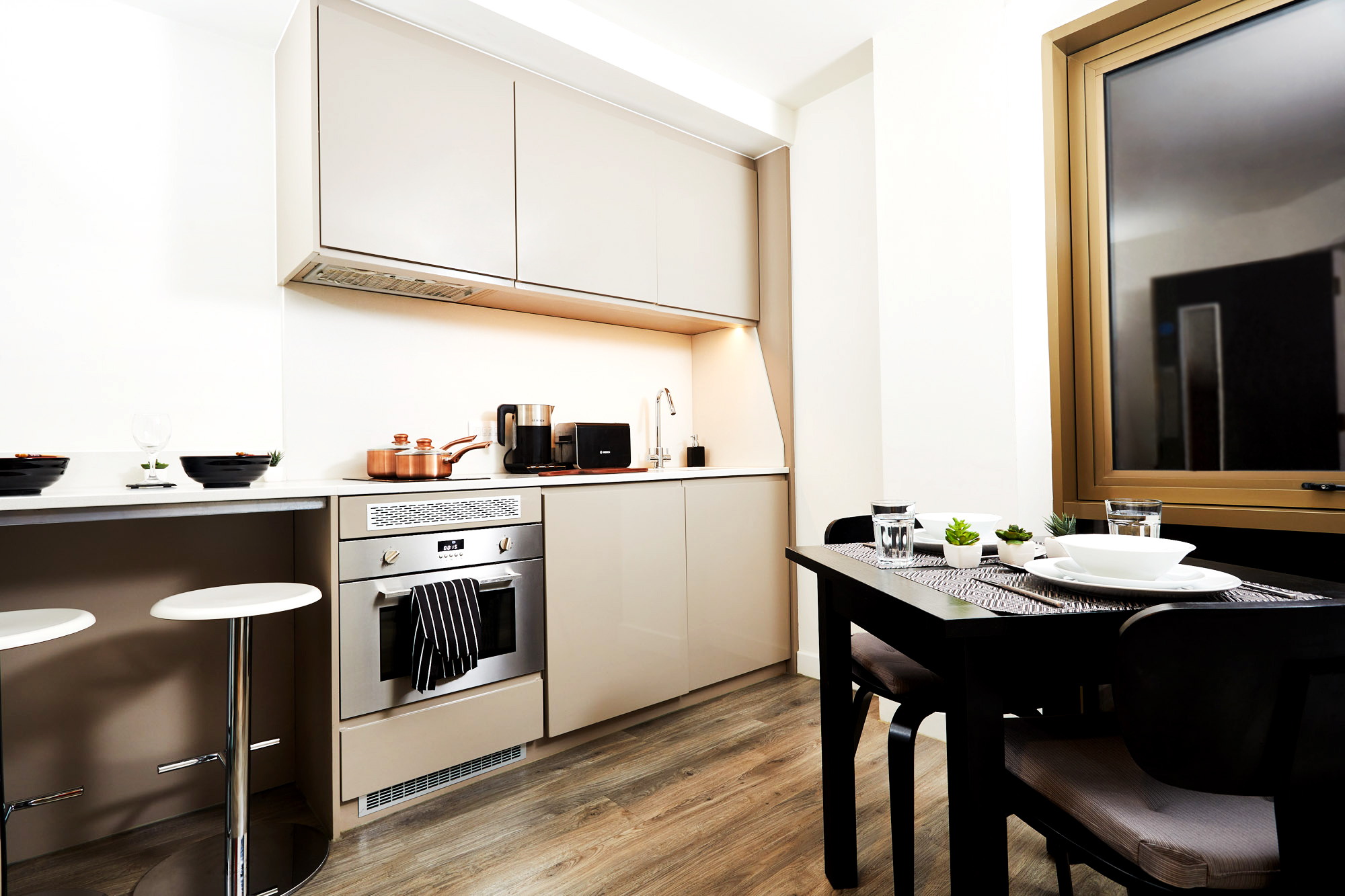 A modern kitchen with cream cabinets, a built-in oven, and a stovetop with copper pots. Black bar stools are at a counter. A dining table with place settings for two sits by a large window, and the floor is wooden.