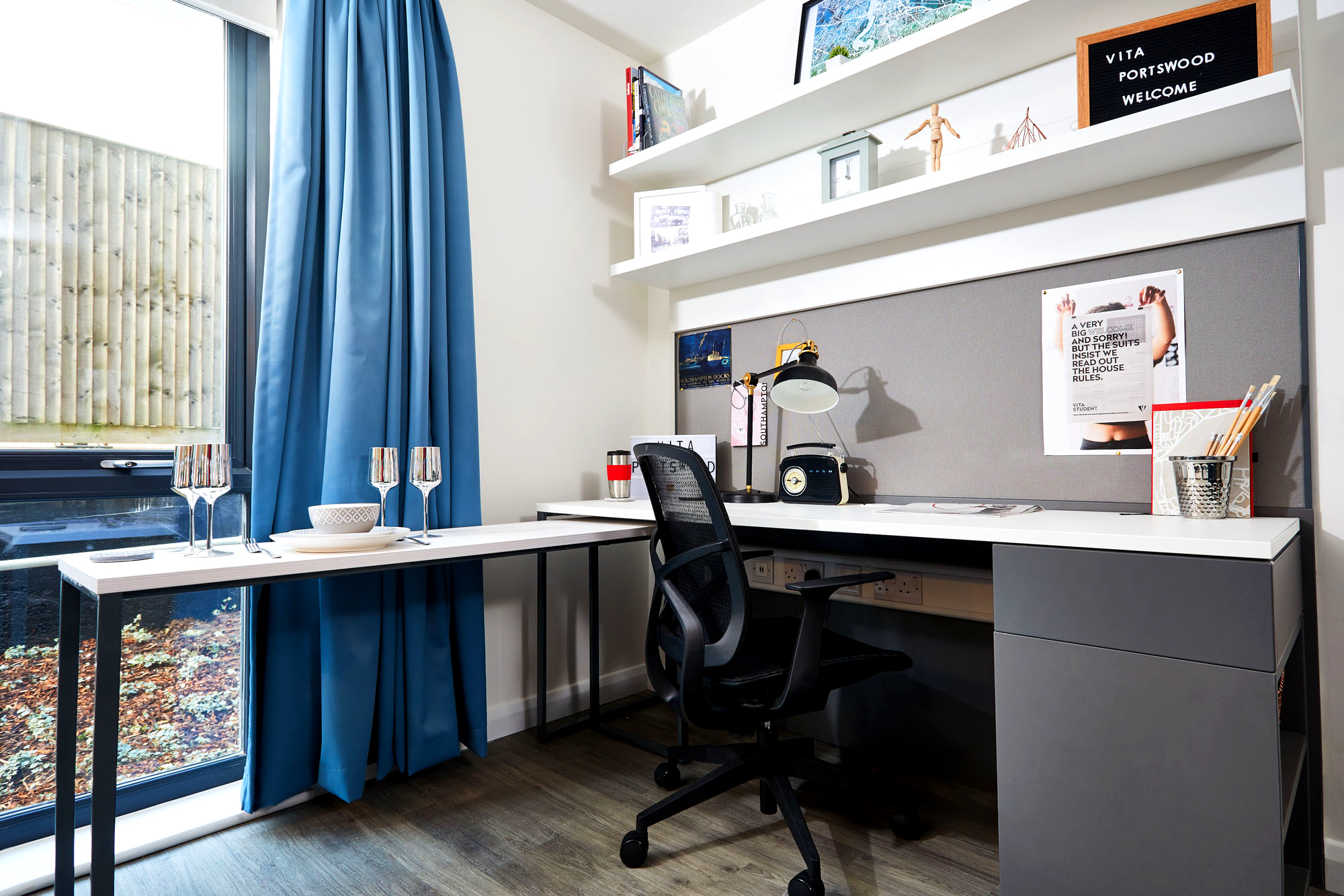 In Portswood House, a modern study space features a corner desk, black chair, and pinboard. Shelves above hold books and decor. A large window with blue curtains bathes the room in natural light. To the side, a small table displays two glasses and a bowl.