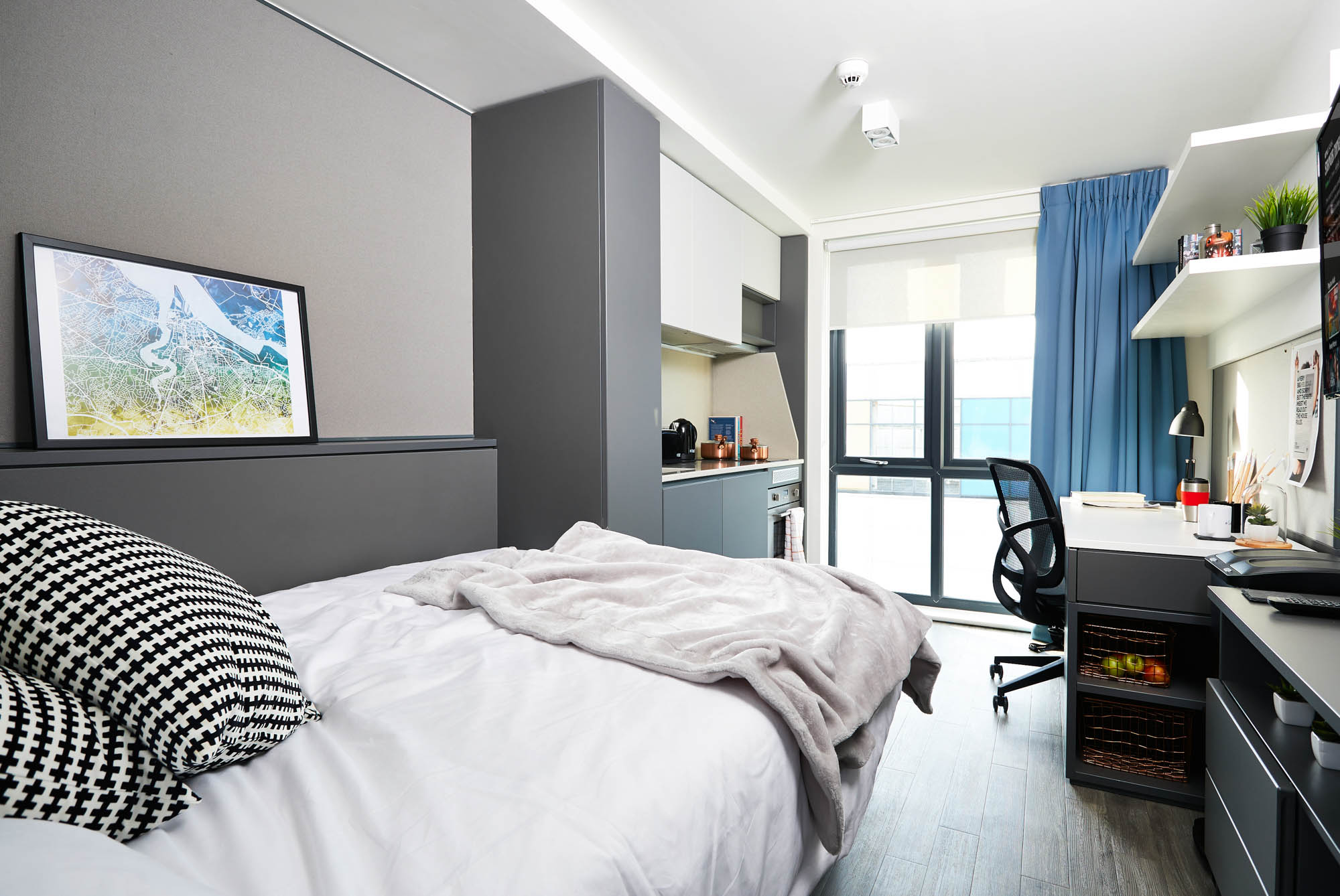 The modern, well-lit bedroom at Portswood House features a neatly made bed with pillows and a blanket. A desk and chair sit by the window, while shelves hold books and decor. A framed map adorns the wall, and blue curtains enhance the room's minimalist design.