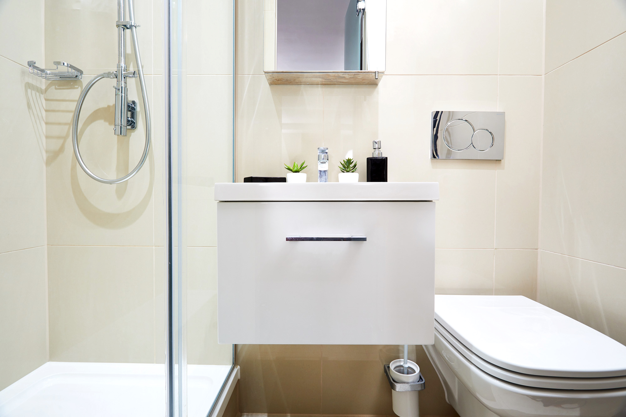 This modern bathroom, perfect for student accommodation, features a glass shower enclosure on the left and a white vanity with a mirror above. Small potted plants add a touch of greenery, while the wall-mounted toilet sits on the right against sleek, neutral-toned tiles.