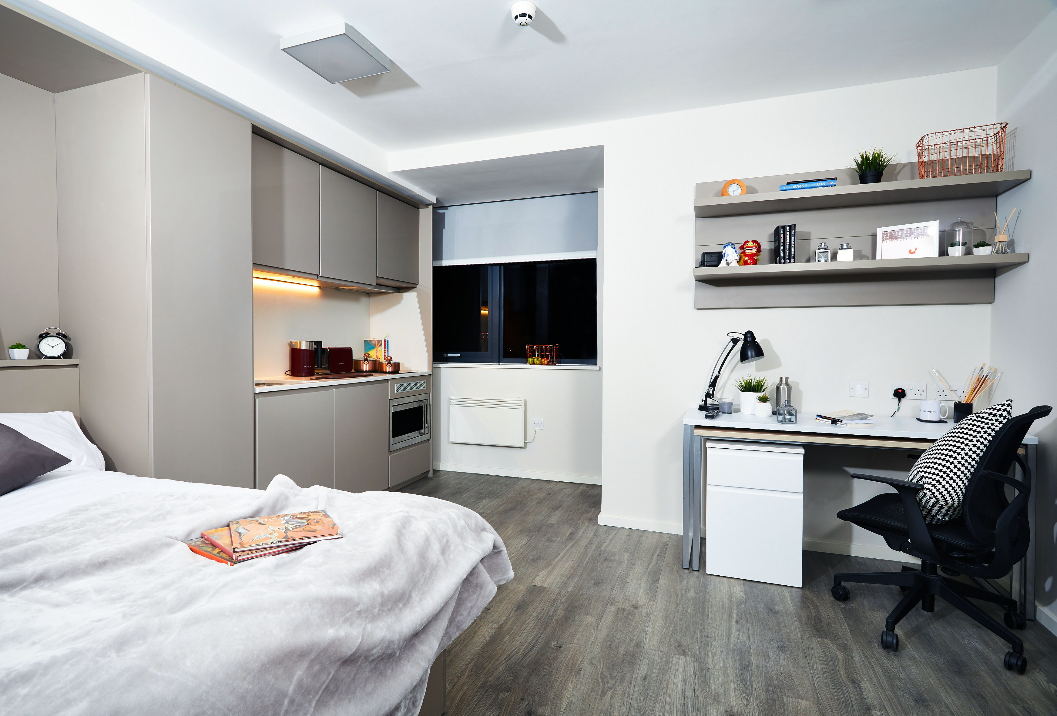 Modern student accommodation featuring a dorm room with a bed, kitchenette, and study area. The bed boasts a gray blanket and book, while the kitchenette offers cabinets and appliances. A desk with a lamp, chair, and shelves adorned with books and decor completes the space. A window with closed blinds lies in the background.