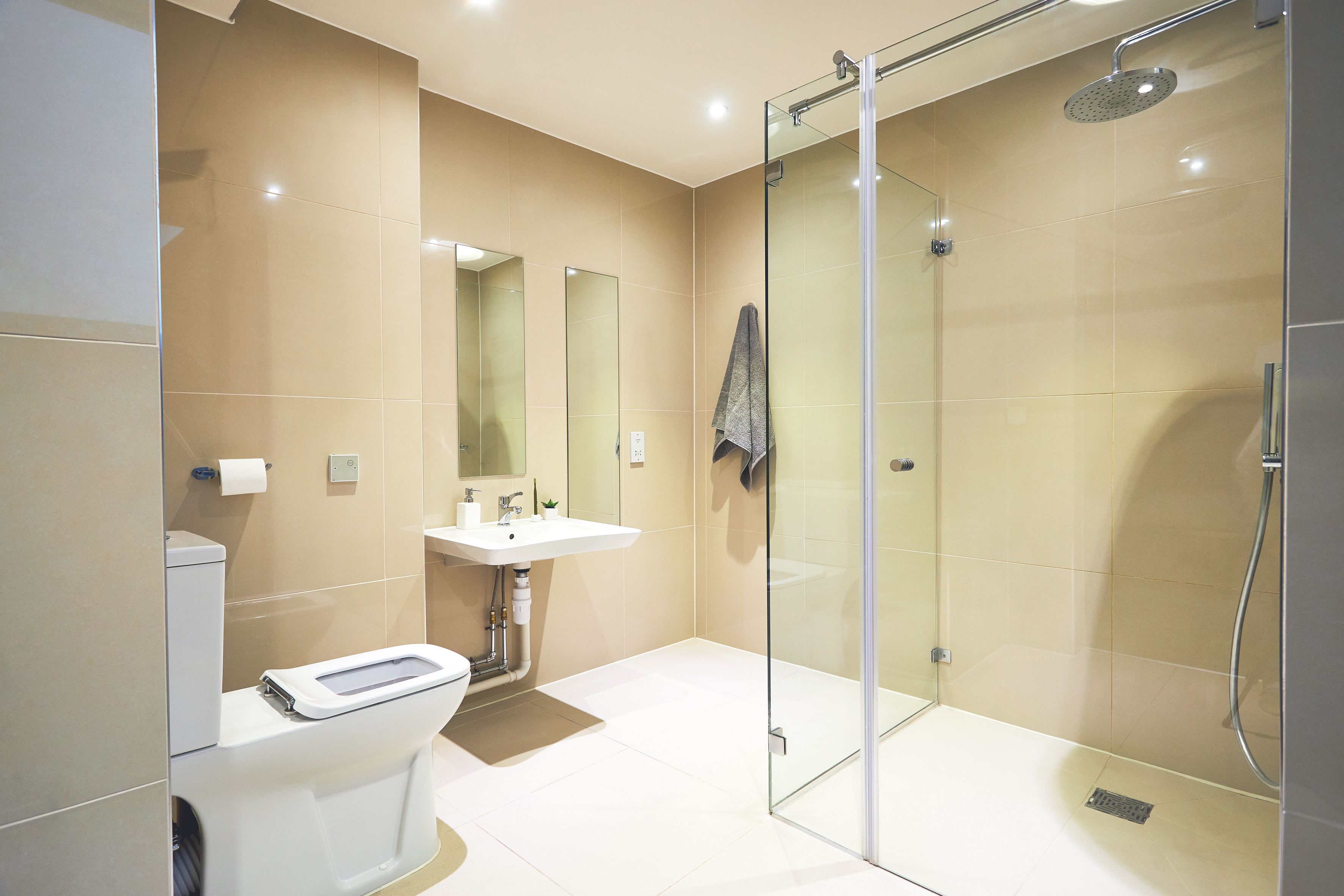 A modern bathroom in a student accommodation features beige tiles, a glass shower enclosure, and a wall-mounted showerhead with a towel on the door. A white toilet and sink with a rectangular mirror and wall-mounted faucet are on the left, while ceiling lights brighten the space.