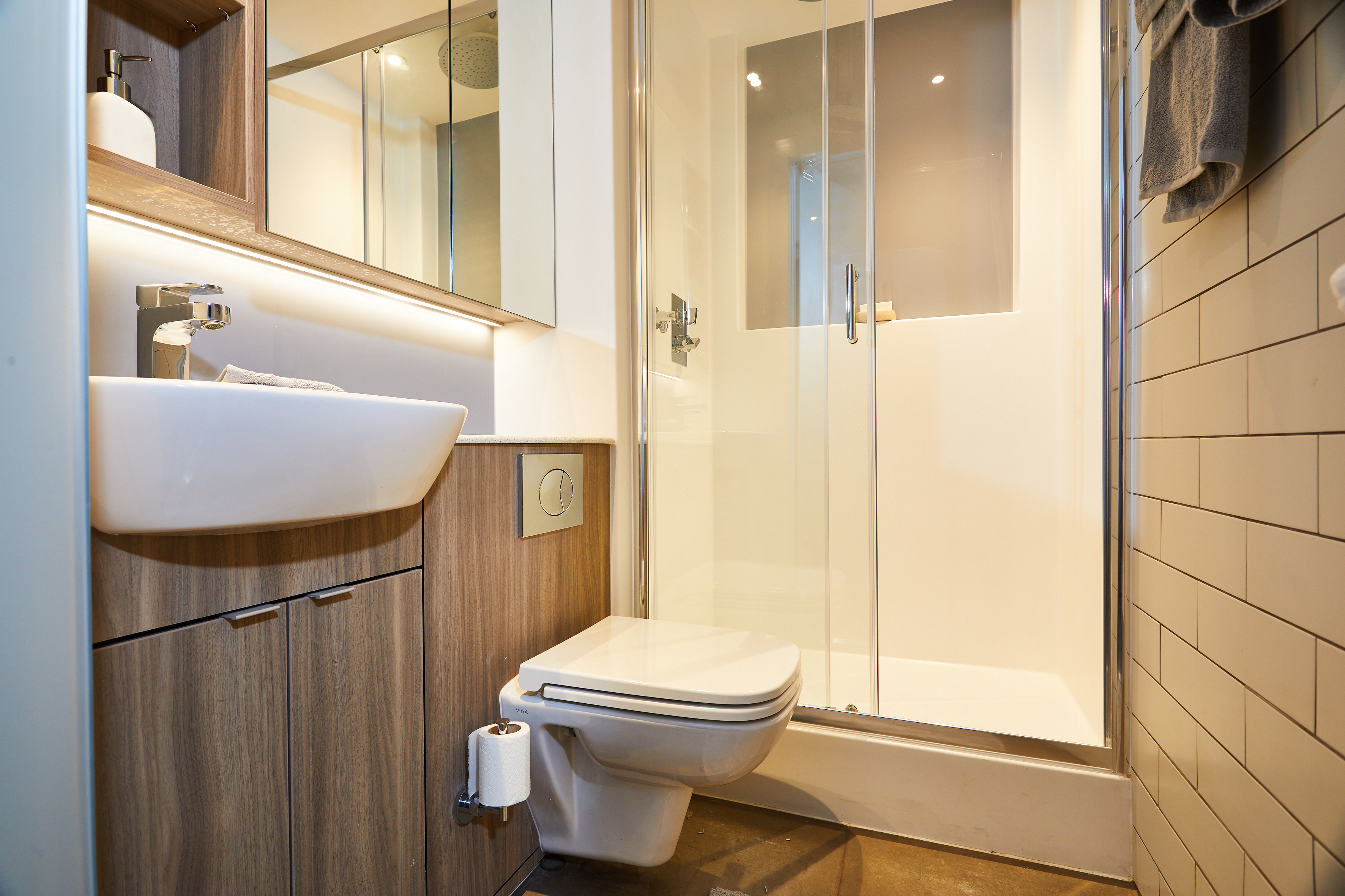The modern bathroom at Portswood House features a wall-mounted sink and toilet, wooden cabinets, and a glass-enclosed shower. White tiled walls and soft lighting enhance its clean, minimalist design. A toilet paper roll sits conveniently on a holder beside the toilet.