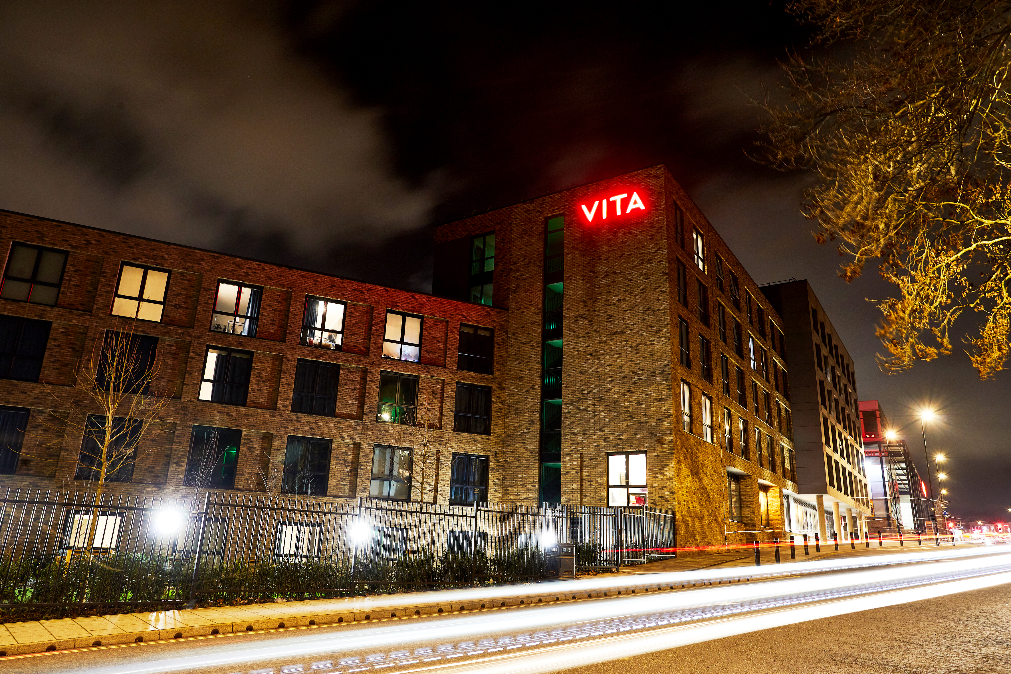 A multi-story brick building at night with "VITA" in red neon letters on the facade. The building is illuminated, and light trails from passing cars are visible on the street in the foreground. Sparse trees are in the scene. Sky is cloudy.