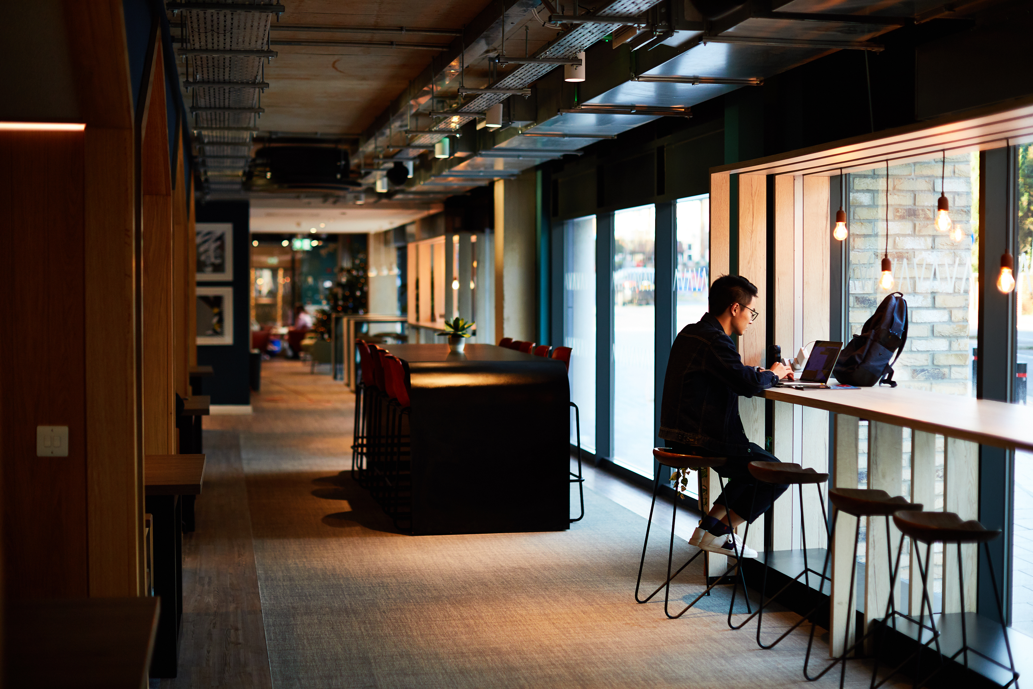 A person sits alone at a long counter beside the floor-to-ceiling windows in Portswood House, a modern, well-lit café. They're working on a laptop, with books and coffee nearby. The spacious interior boasts high ceilings and minimalist design.