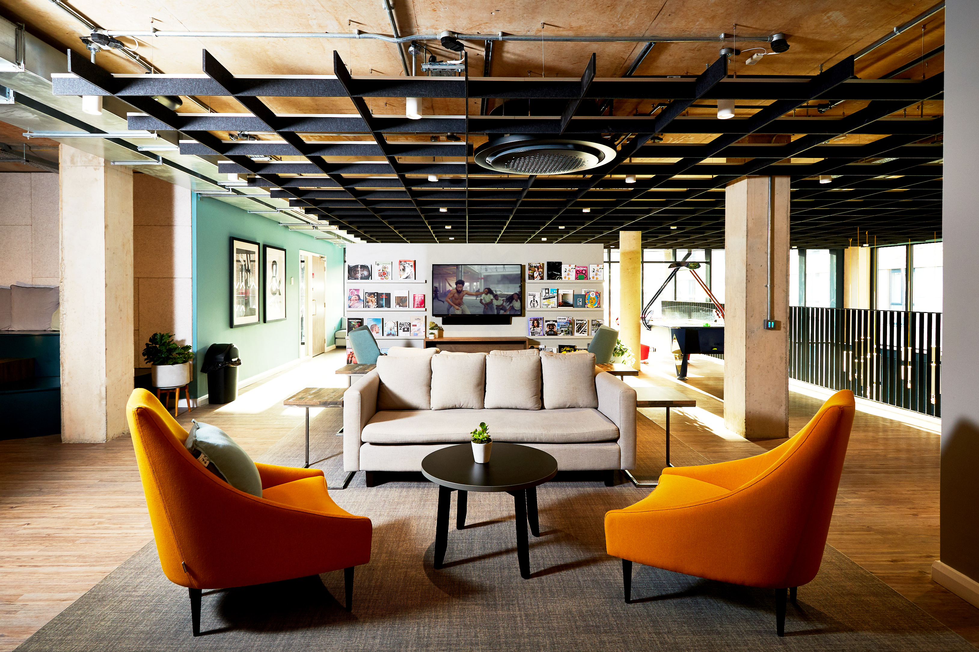 Portswood House offers a modern, well-lit office lounge with two orange chairs and a beige sofa around a black table on a rug. The room features wooden flooring, an exposed ceiling, framed artwork on the walls, and large windows that flood the space with natural light.