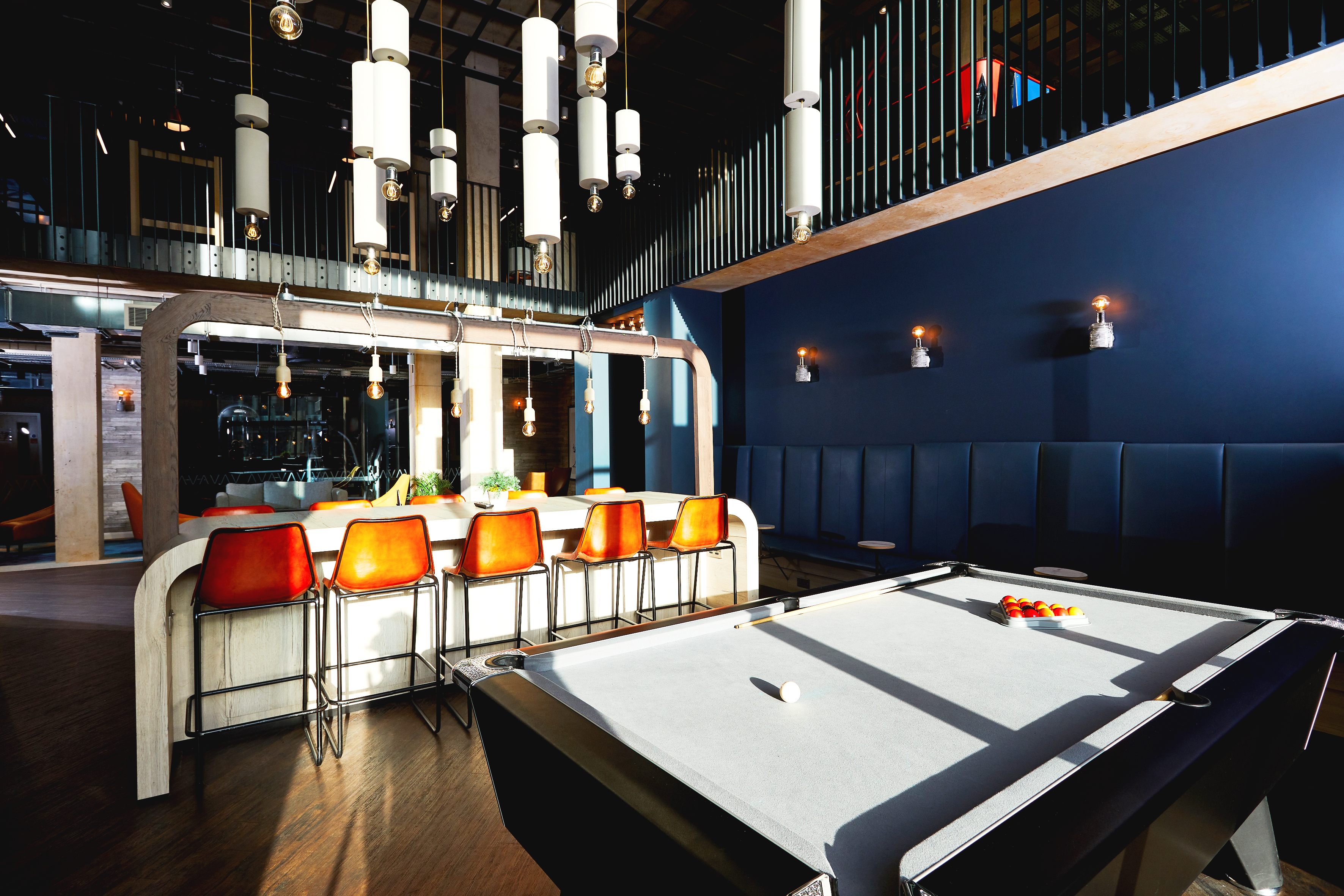 Modern room with a pool table in the foreground, bar with red stools, and contemporary lighting. Dark walls and ceiling feature a mix of wood and metal accents. Natural light streams in, enhancing the sleek atmosphere of this stylish Southampton Property.