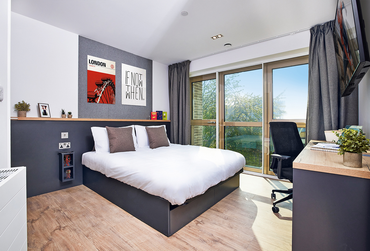 A modern Lewisham bedroom with a large bed, white bedding, and gray pillows. A desk with a chair, monitor, and potted plant sits by floor-to-ceiling windows. A "LONDON" poster and decor items adorn the walls above the bed. Light wood flooring enhances the airy space.