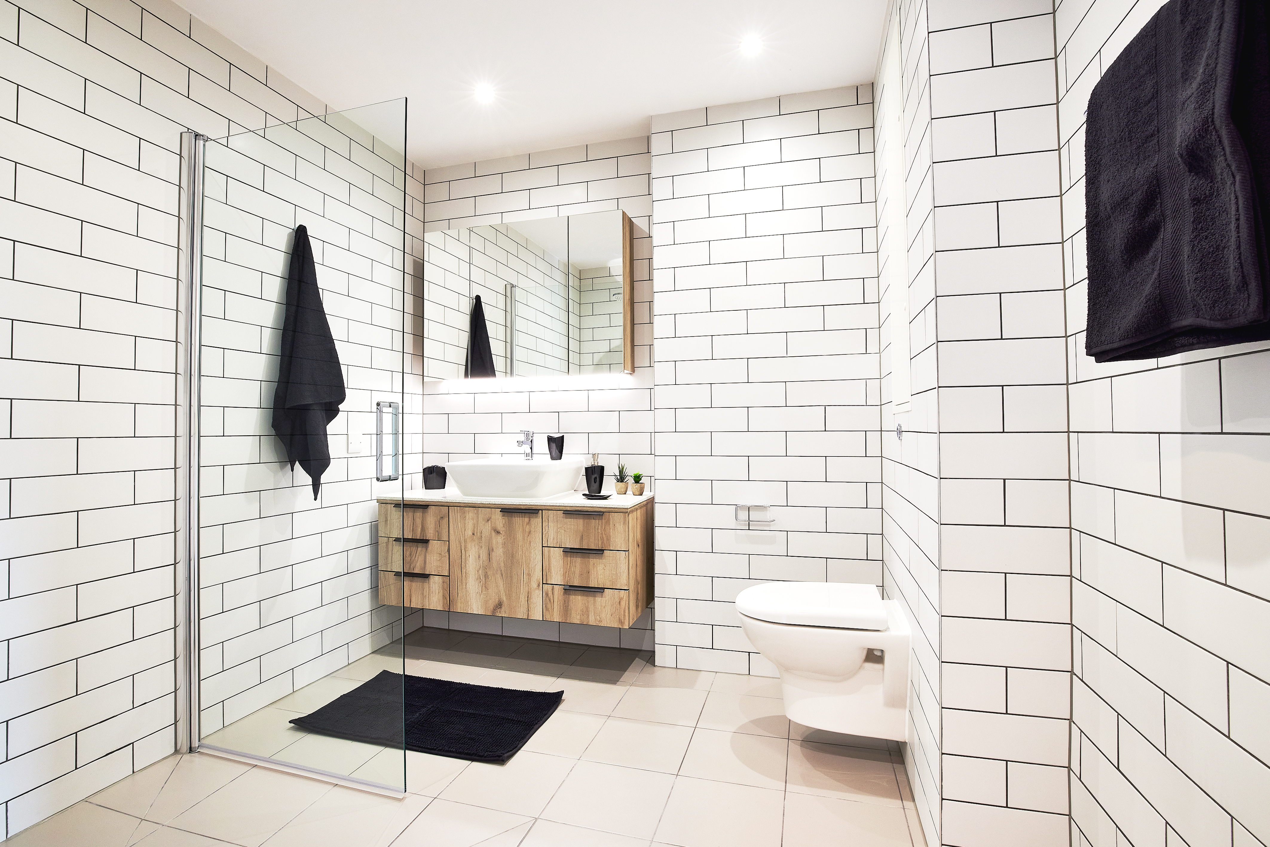 This modern bathroom at Lewisham Exchange features white subway tiles, a sleek glass shower, and a wooden vanity with a stylish sink. A wall-mounted mirror reflects the space's elegance, while black towels and a rug add contrast. Ceiling lights beautifully illuminate the white toilet on the right.