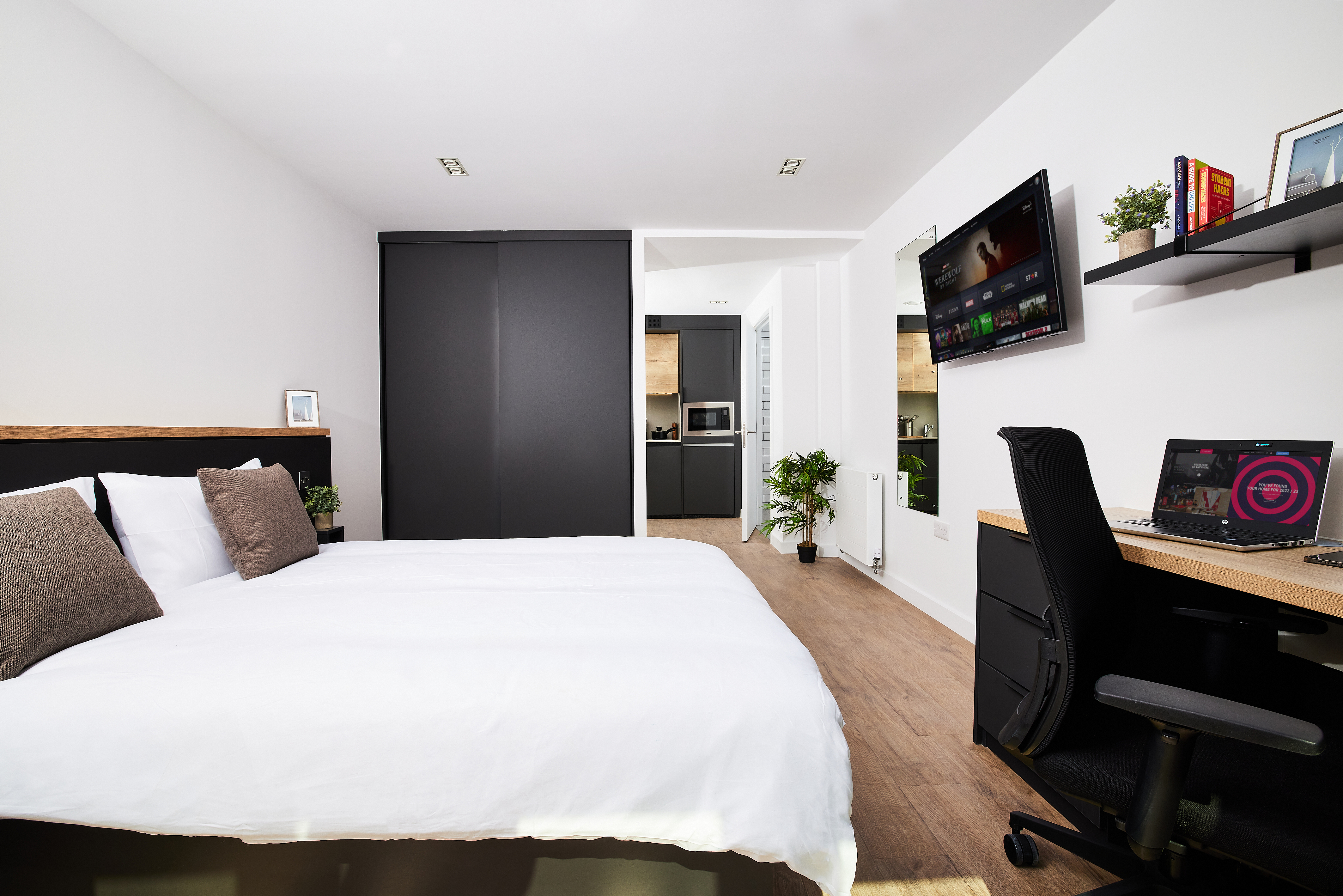 This modern Lewisham bedroom features a large bed with white bedding and dark pillows. A sleek black wardrobe stands against the wall, while a flat-screen TV is mounted opposite. A desk with a computer, shelves, and small plants completes the space, all set on elegant wooden flooring.