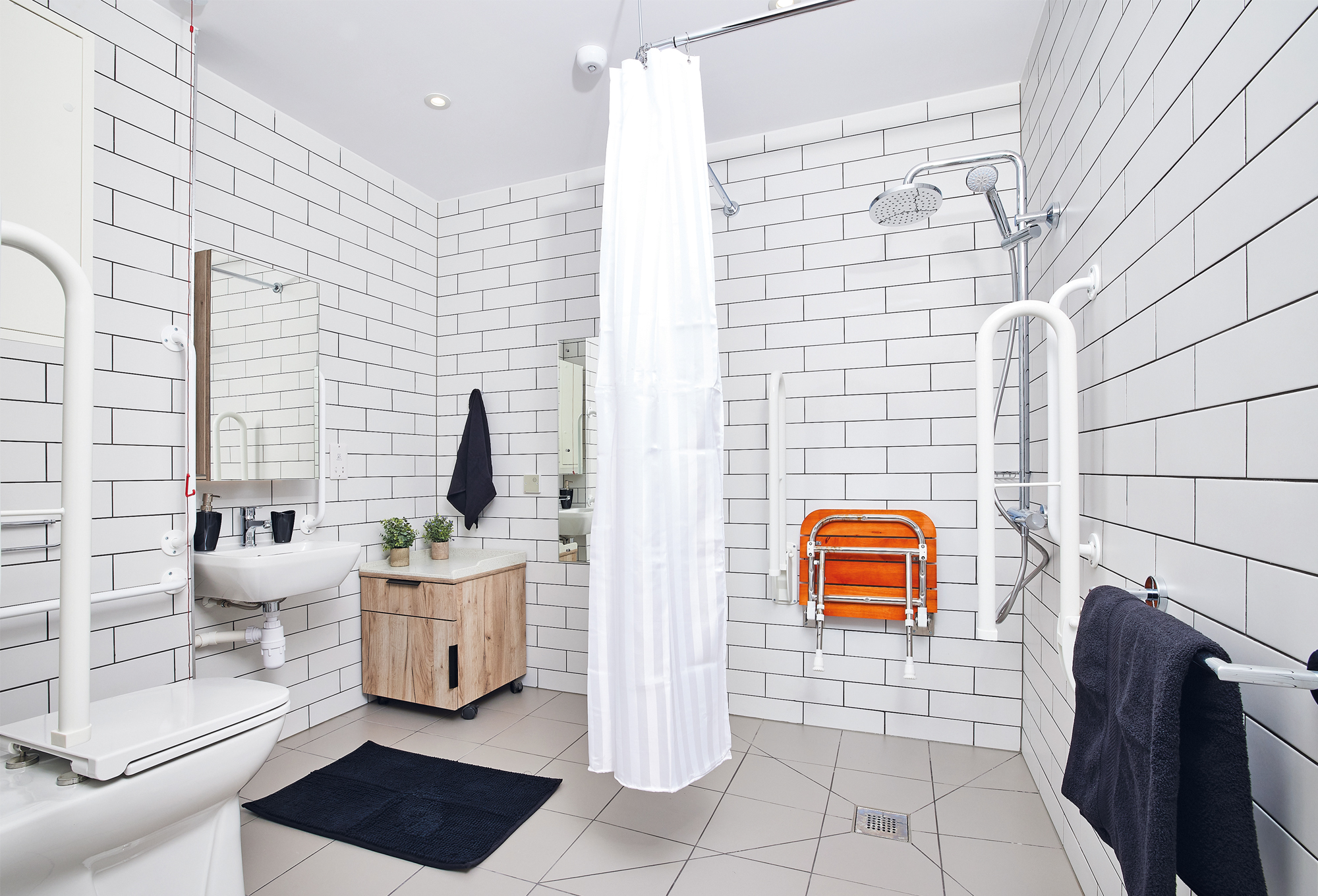 The Lewisham Exchange bathroom boasts modern accessibility features, including white subway tiles, a wall-mounted sink, and a shower with a white curtain and safety grab bars. A foldable orange seat complements the black mat on the floor, ensuring both style and functionality.