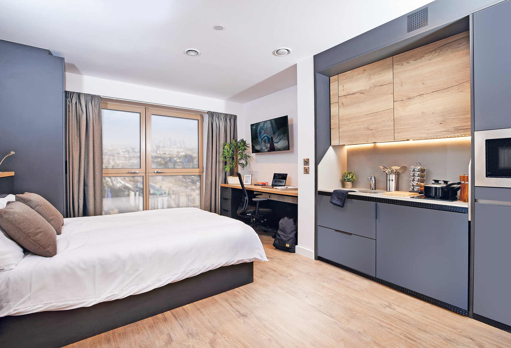 Modern studio apartment in Lewisham with a neatly made bed, large window offering a city view, and a work desk with a computer. The kitchen area features sleek cabinets, a microwave, and cookware. The wood floors enhance the minimalist decor, creating an inviting urban retreat.