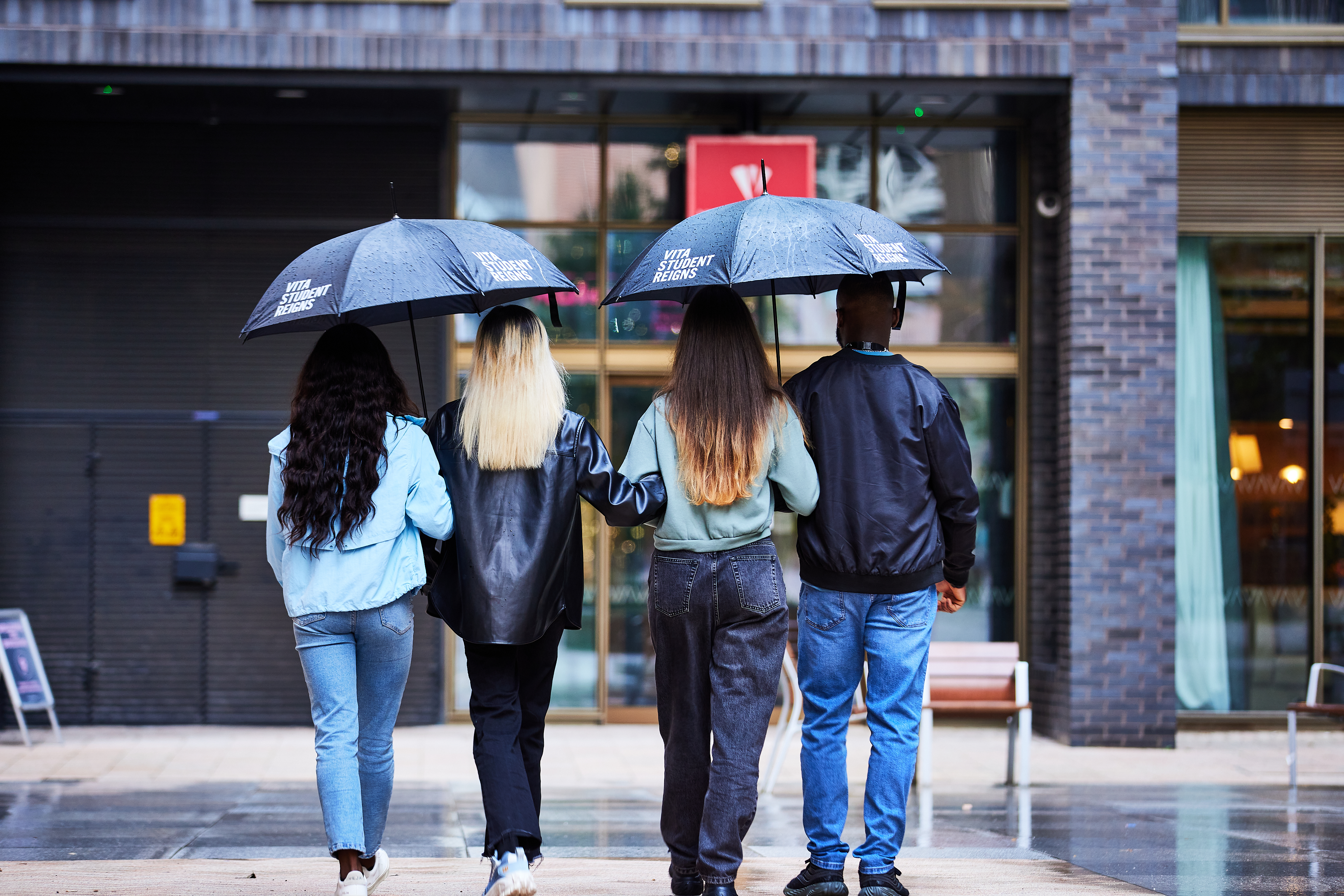 Four people walk under two umbrellas on a rainy street. The group, sporting various hairstyles and casual jackets, strides towards a building with large windows. Their attire, consisting of jeans and other casual wear, reflects a relaxed UK style as they head away from the camera.