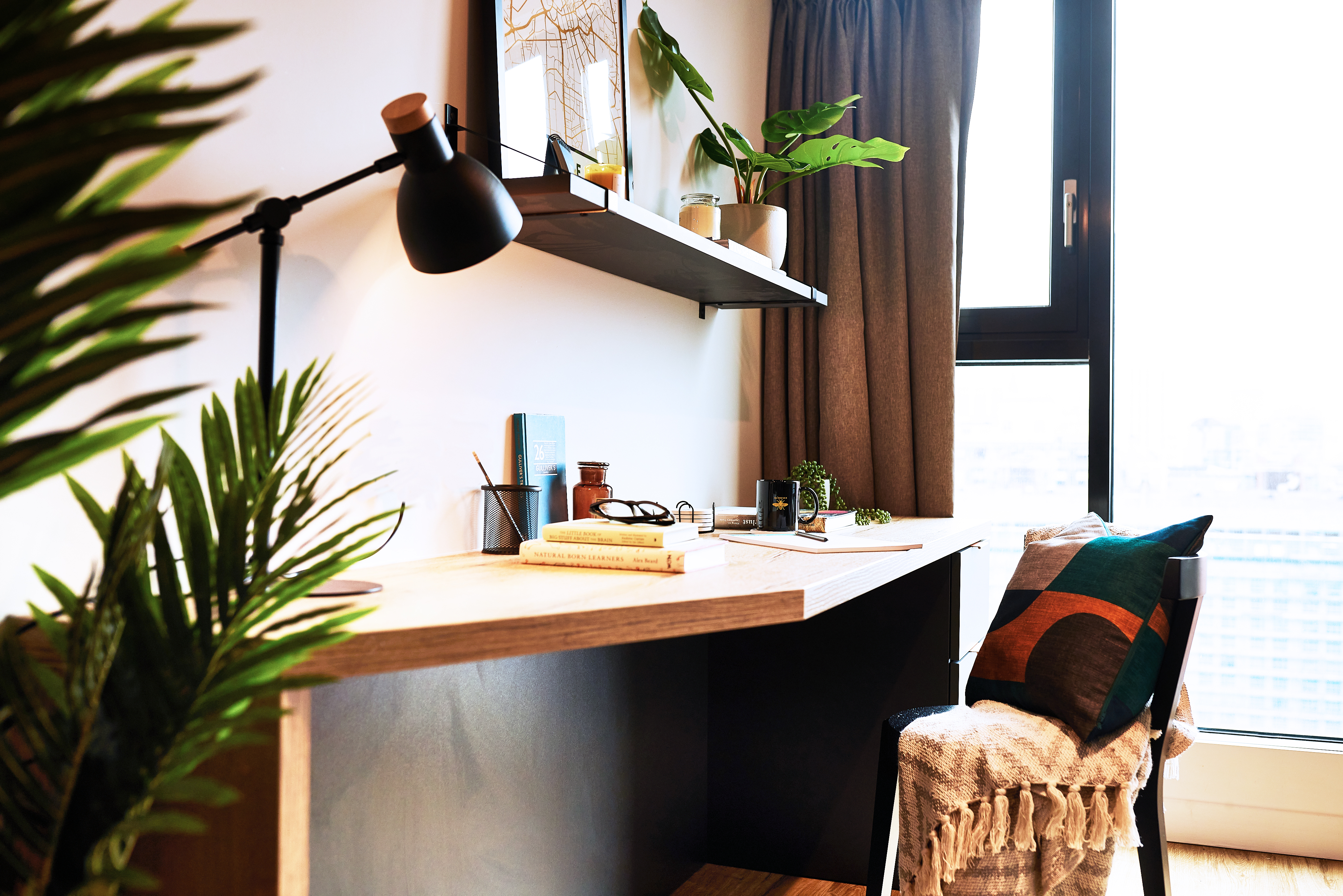A cozy workspace features a wooden desk with a black lamp, books, and a circular plant pot among the decorative items. A square shelf adorned with framed art and a potted plant is above. Nearby, a chair with a patterned pillow and throw sits next to a large window with curtains.
