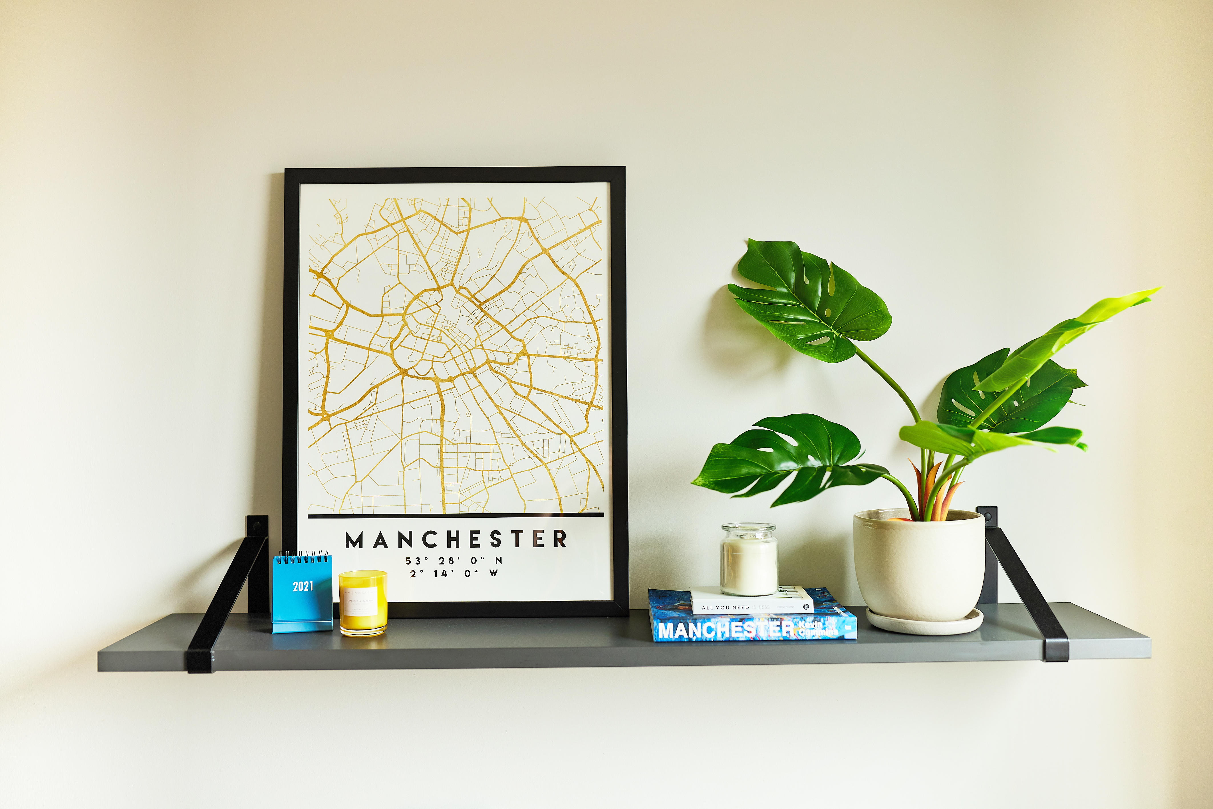 A shelf against a white wall holds a framed map of Manchester, a potted plant with large green leaves, three small colorful books, two candles, and a square picture frame.