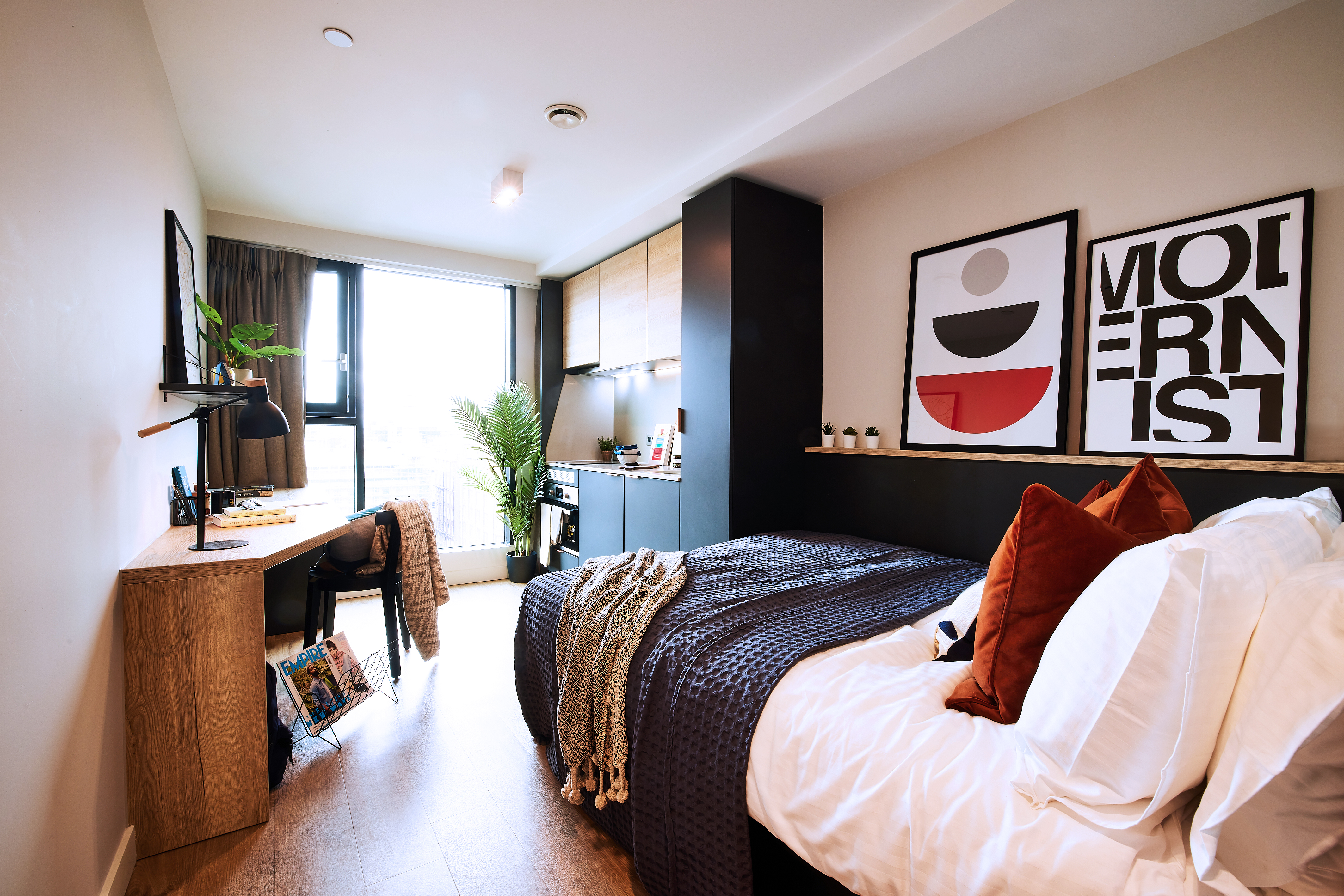 A modern studio apartment features a bed with cushions, a square desk with a computer, and artwork on the walls. A window lets in natural light, while a potted plant adds greenery. The decor is minimalistic with wood accents and a neutral color palette.