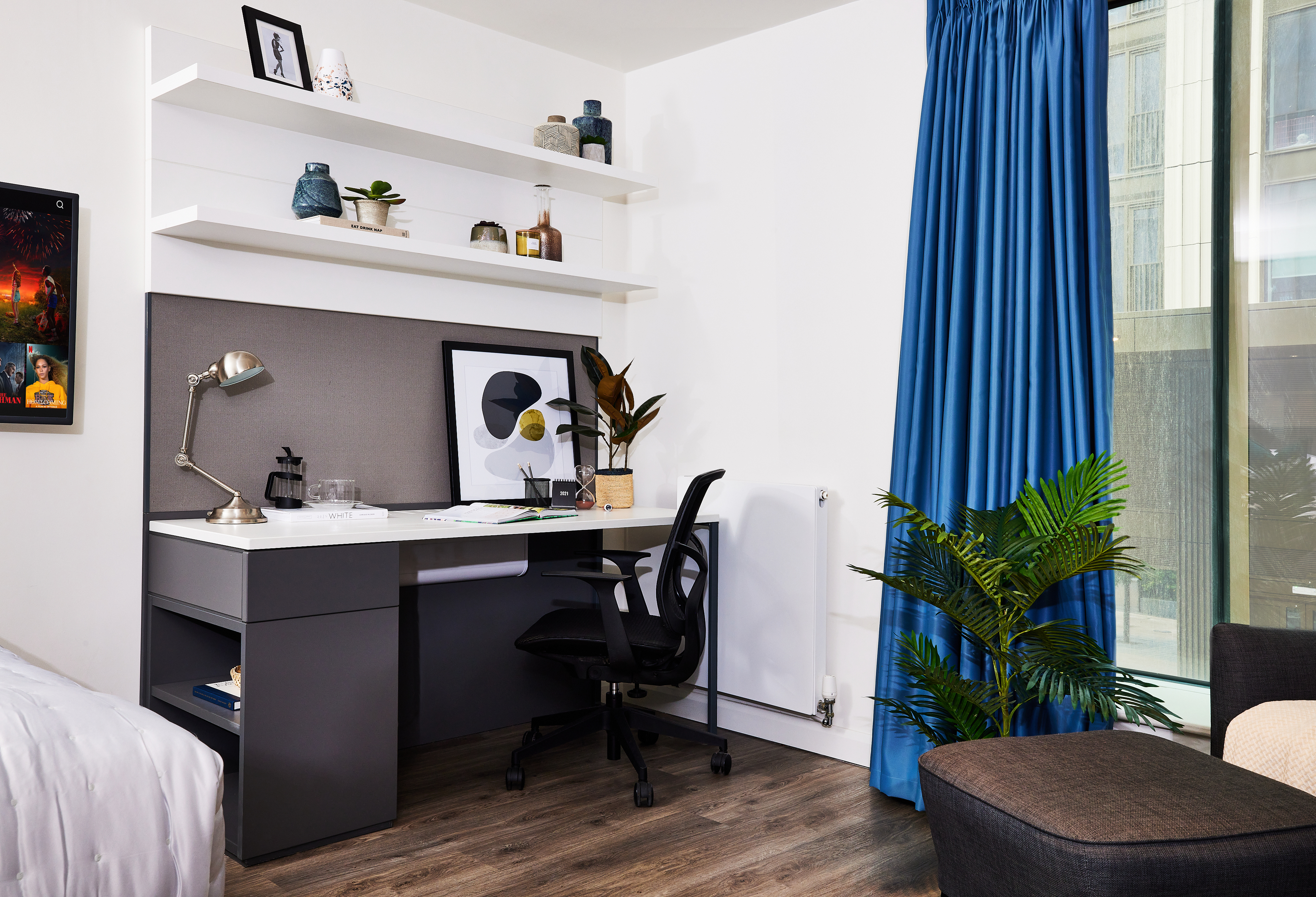 A modern room features a study desk and chair against a wall, complete with a lamp, plants, and decorative items. Circle motifs adorn the curtains framing the large window on the right. Above the desk, shelves hold vases and framed art. A bed is partially visible on the left.