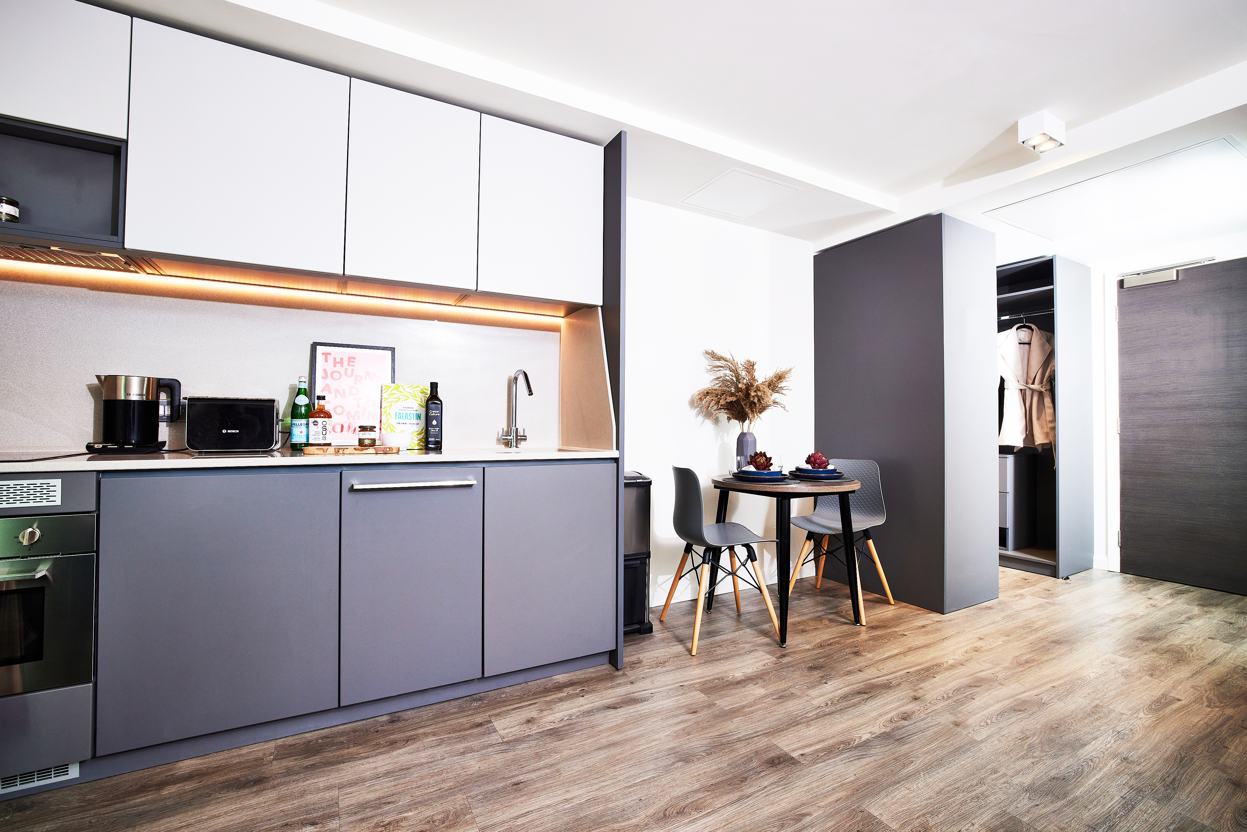 Modern kitchen with gray cabinets and light countertops. Includes a sink, stove, oven, and appliances. A small dining area with a square table set with fruit and two chairs. Coat rack visible in an open wardrobe. Light wood flooring throughout.