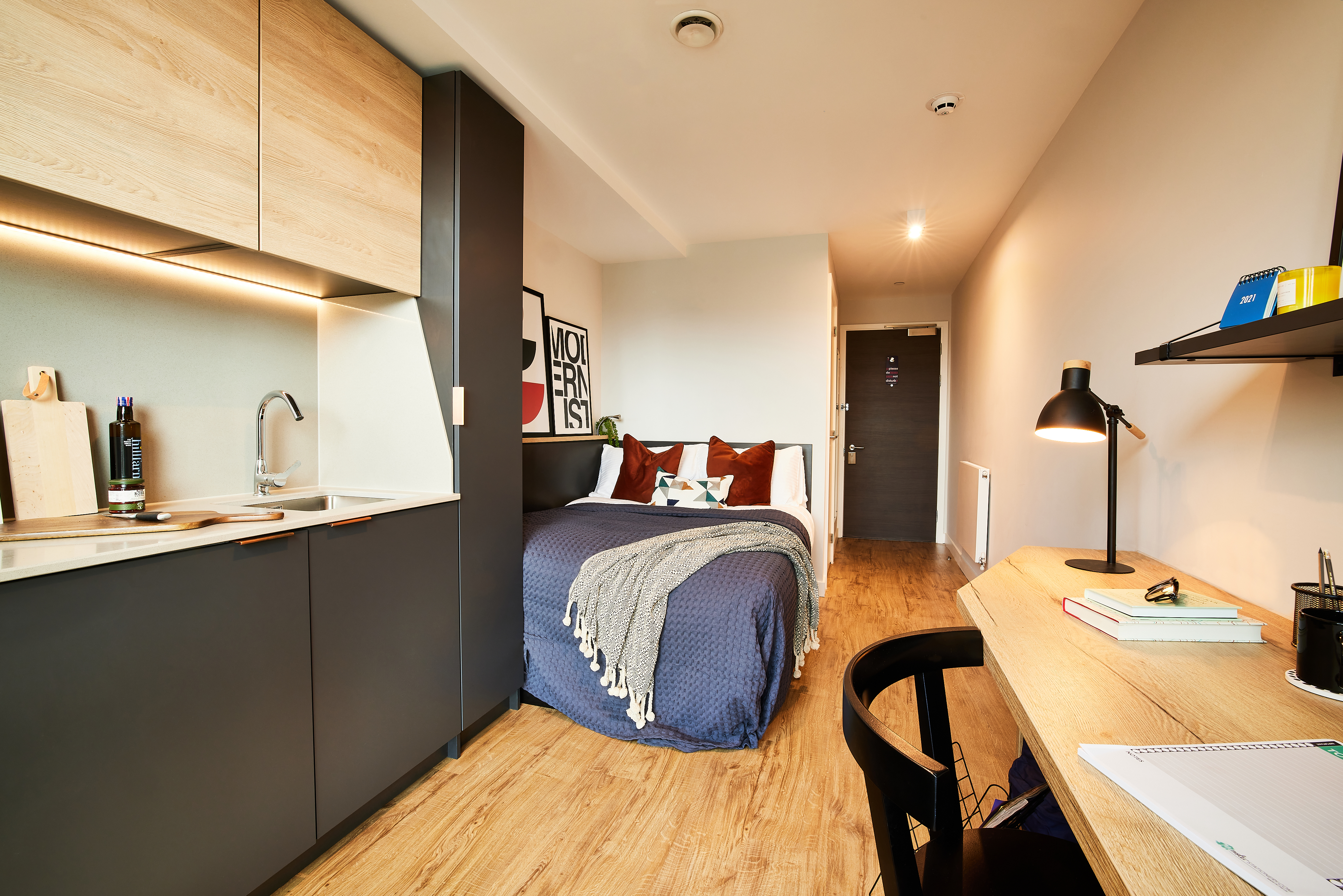 Compact studio apartment with a modern kitchen on the left, featuring a small sink and cabinets. A cozy bed with pillows and a blanket is in the center. A wooden desk with a lamp and books forms a perfect square on the right, and the room has elegantly finished wooden flooring.