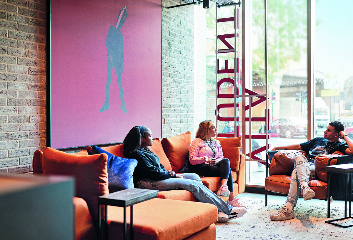 Three people sit on orange sofas in a modern room with a large window. A pink art piece with a silhouette hangs on the brick wall. The space is bright, welcoming, and features wooden tables and a patterned floor rug.